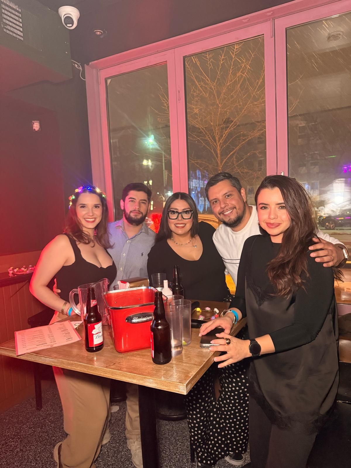 A group of friends enjoying drinks and socializing at a bar with pink lighting and wooden tables.