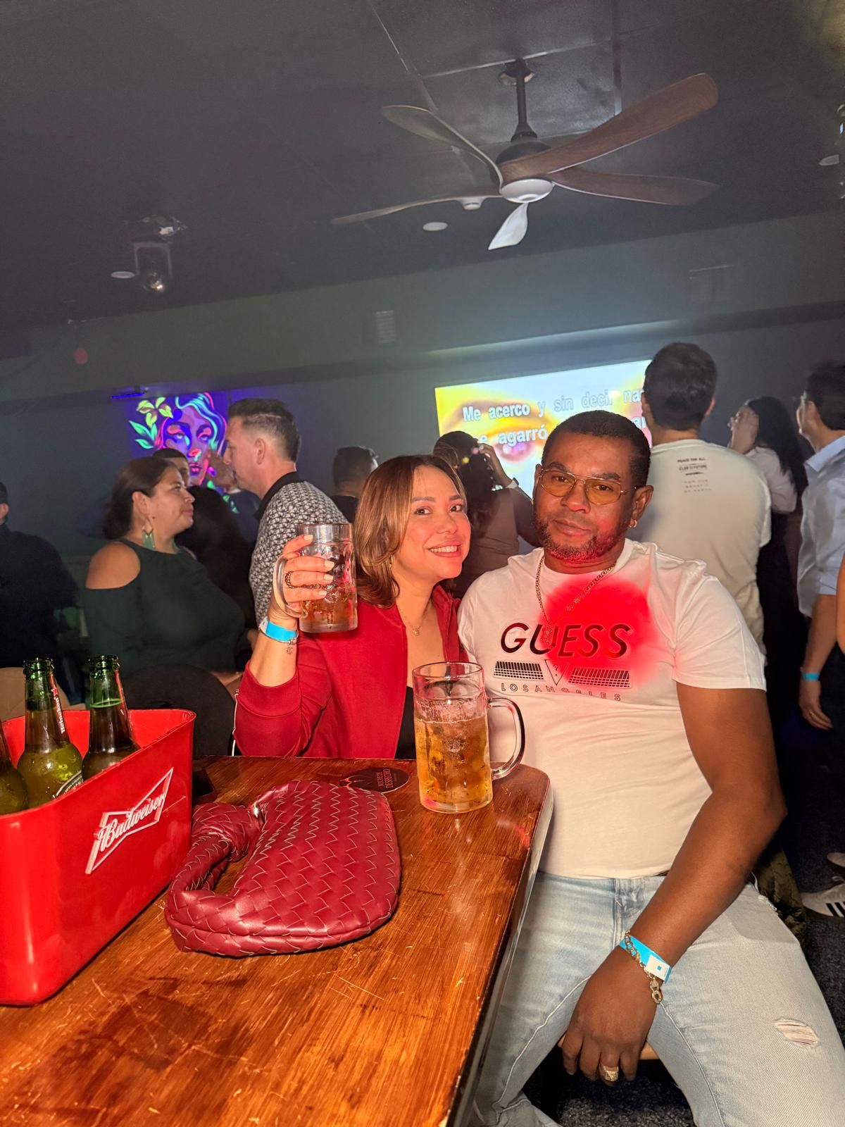 People enjoying drinks and socializing at a bar with red Budweiser coolers and a wooden table in a dimly lit setting.