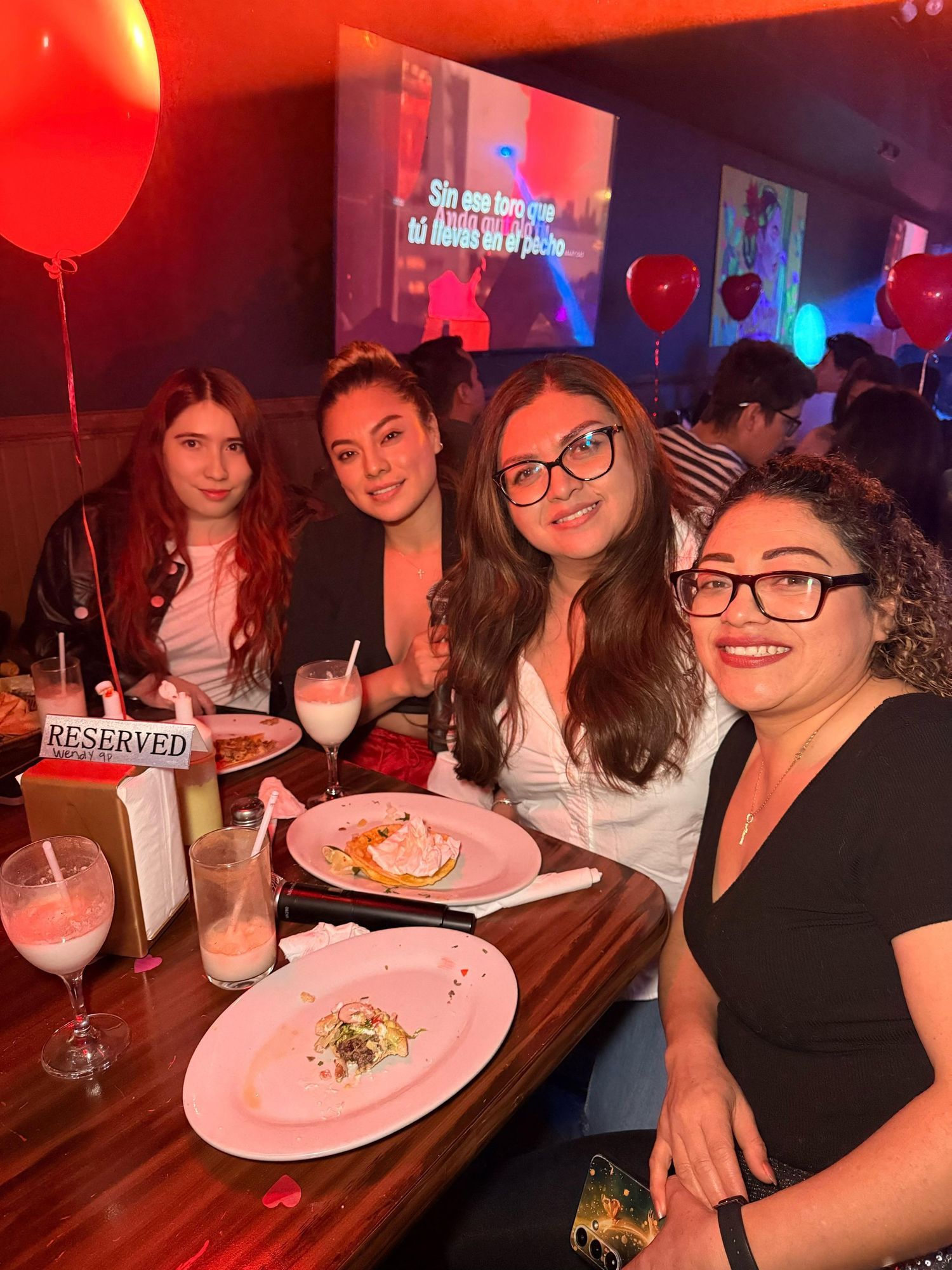 Friends gather around a table with pink plates and red balloons at a restaurant celebration.