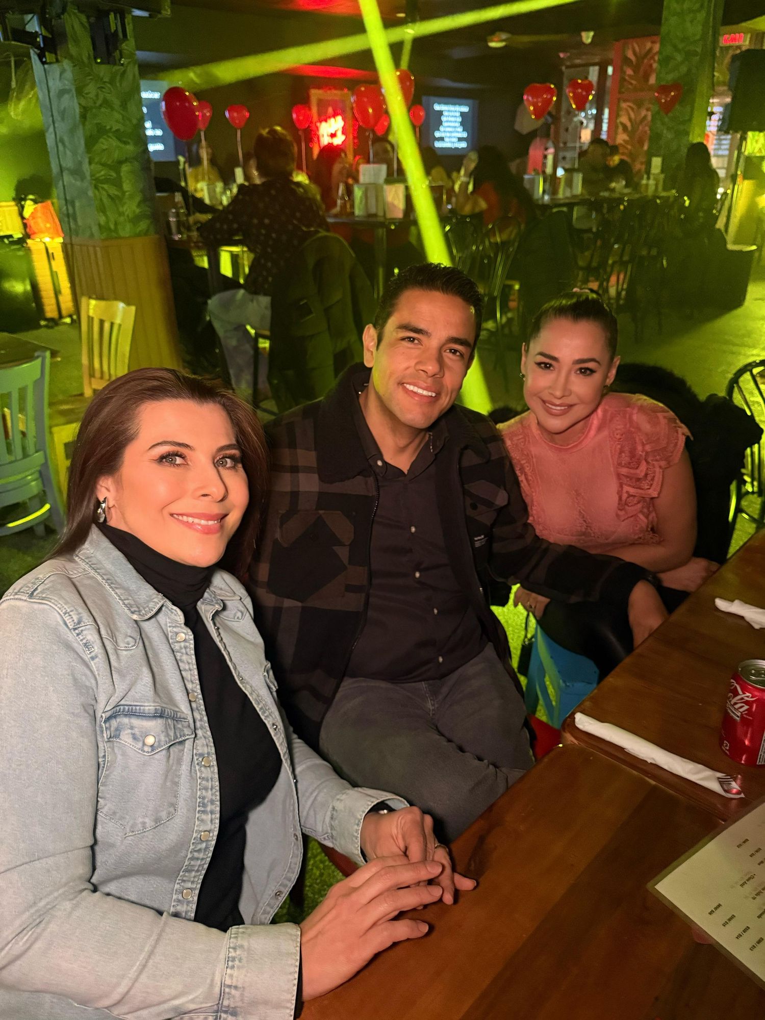 Three people smiling at restaurant table with colorful lighting.