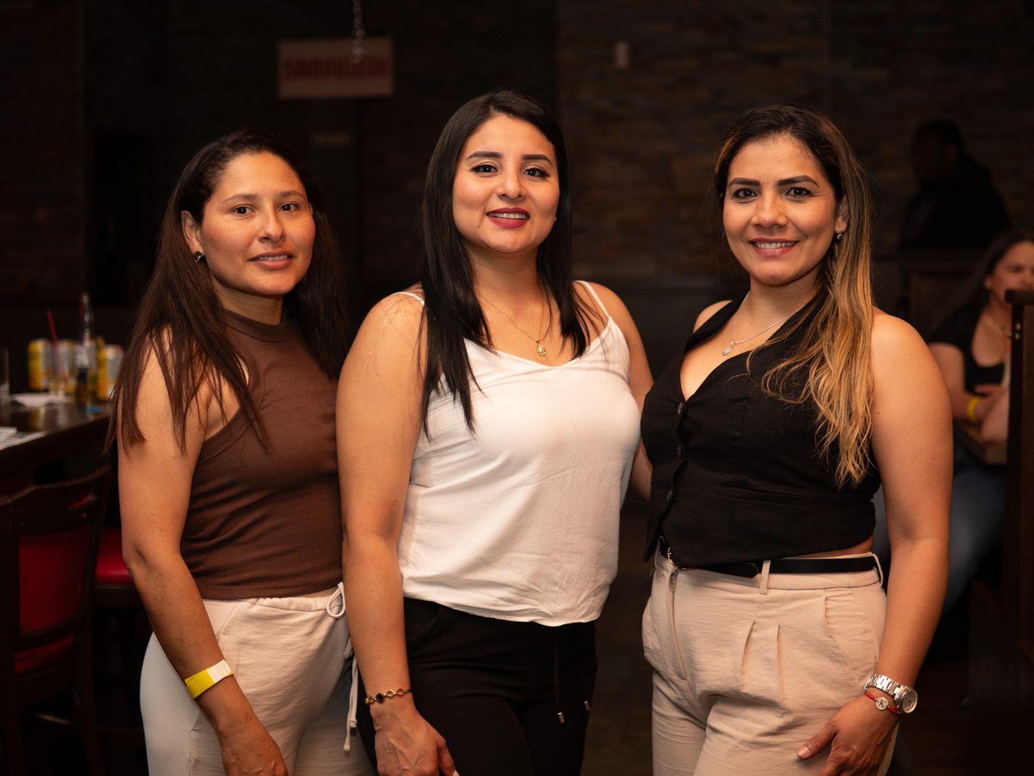Three young women smiling and posing together at a bar or nightclub venue.