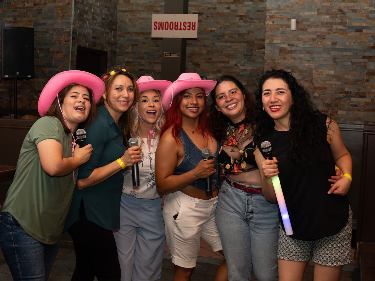 Group of women in pink cowboy hats singing karaoke at a party, holding microphones and smiling.