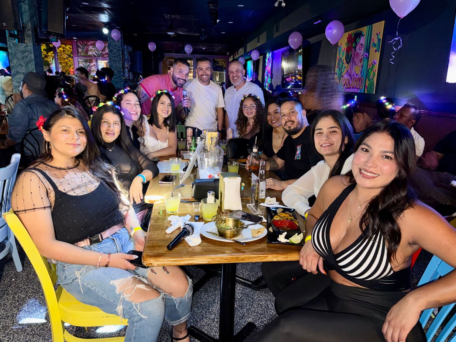 Group of friends smiling and posing at a restaurant table with drinks and food during a festive celebration.