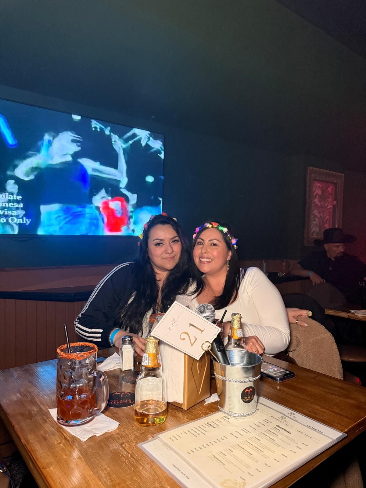 Two patrons sitting at a wooden table in a dark bar with blue lighting and menus visible.