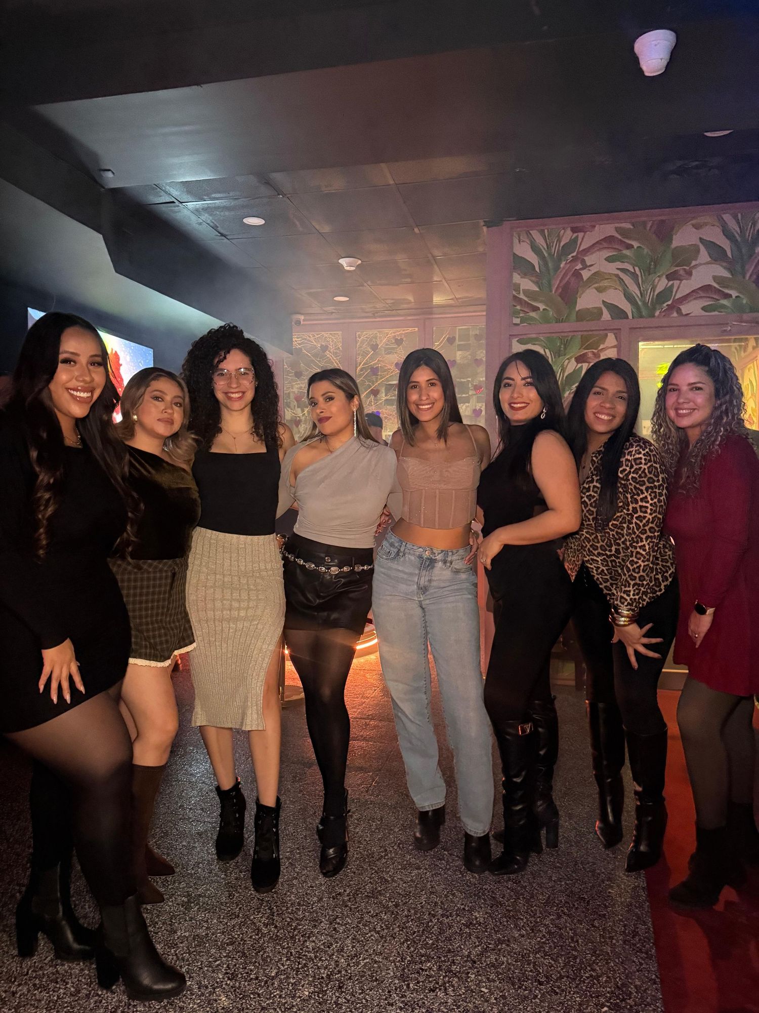 A group of eight women in stylish outfits pose together at a dimly lit venue with decorative wall art.
