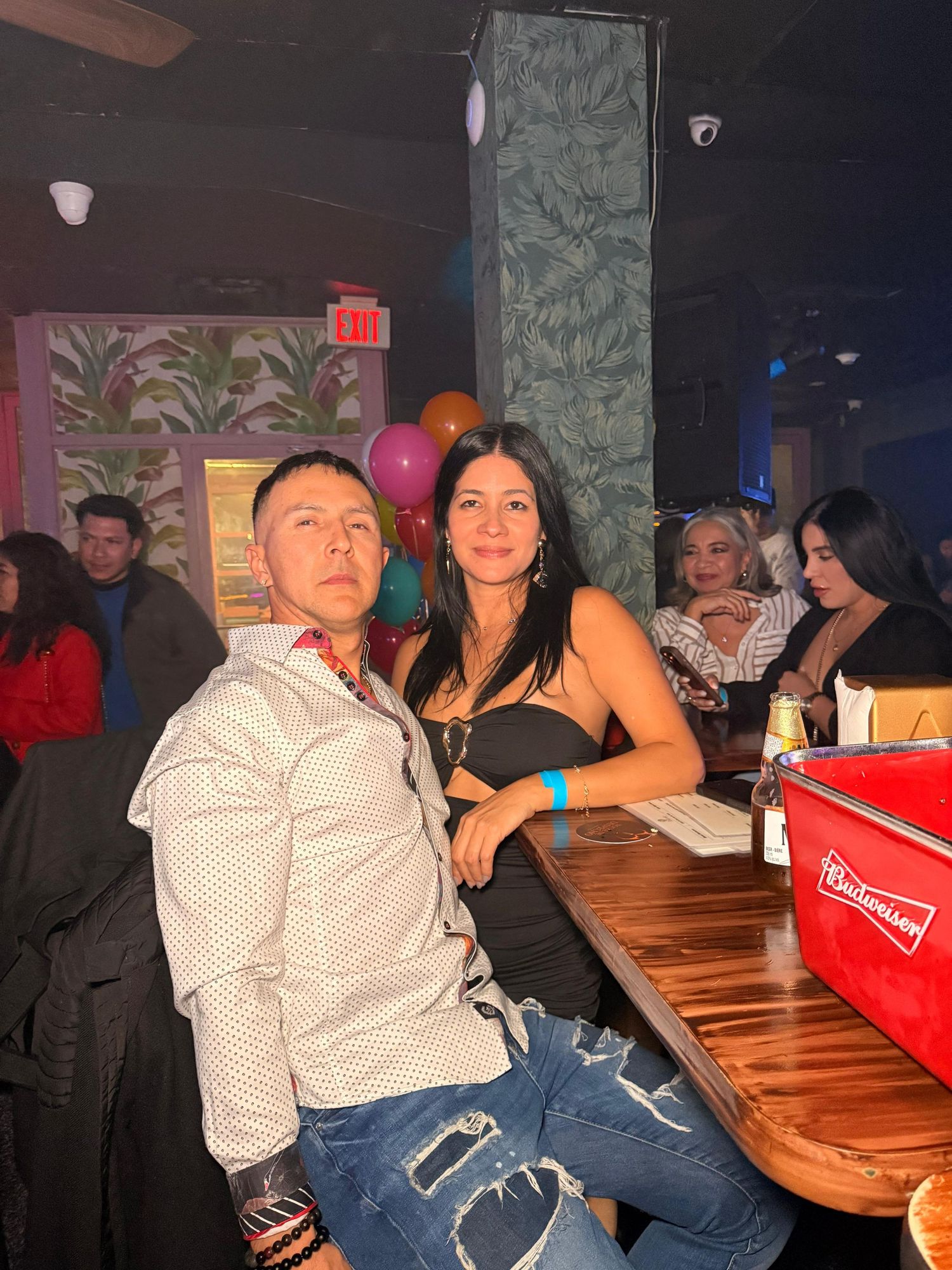 A couple sits at a wooden table in a bar with tropical leaf decor and ambient lighting.