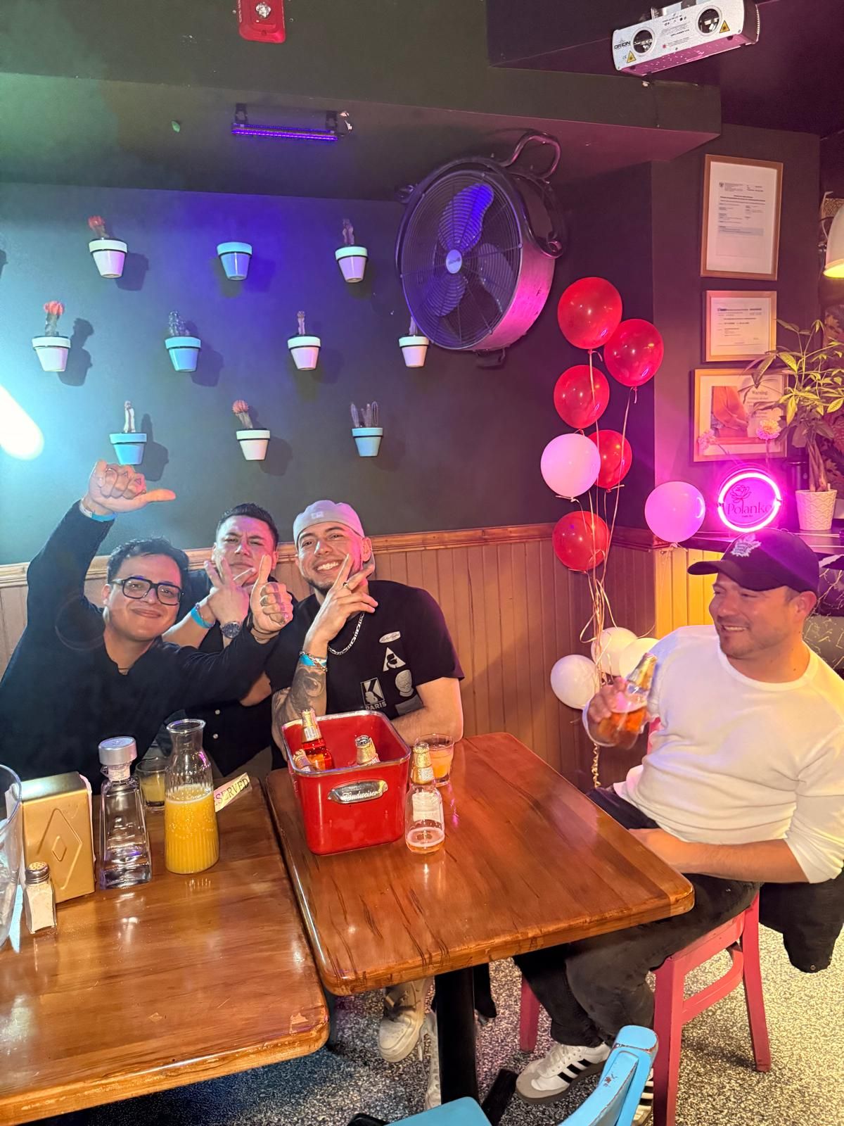 Friends gathered around wooden tables at a dimly lit bar with blue lighting and red balloons for a celebration.