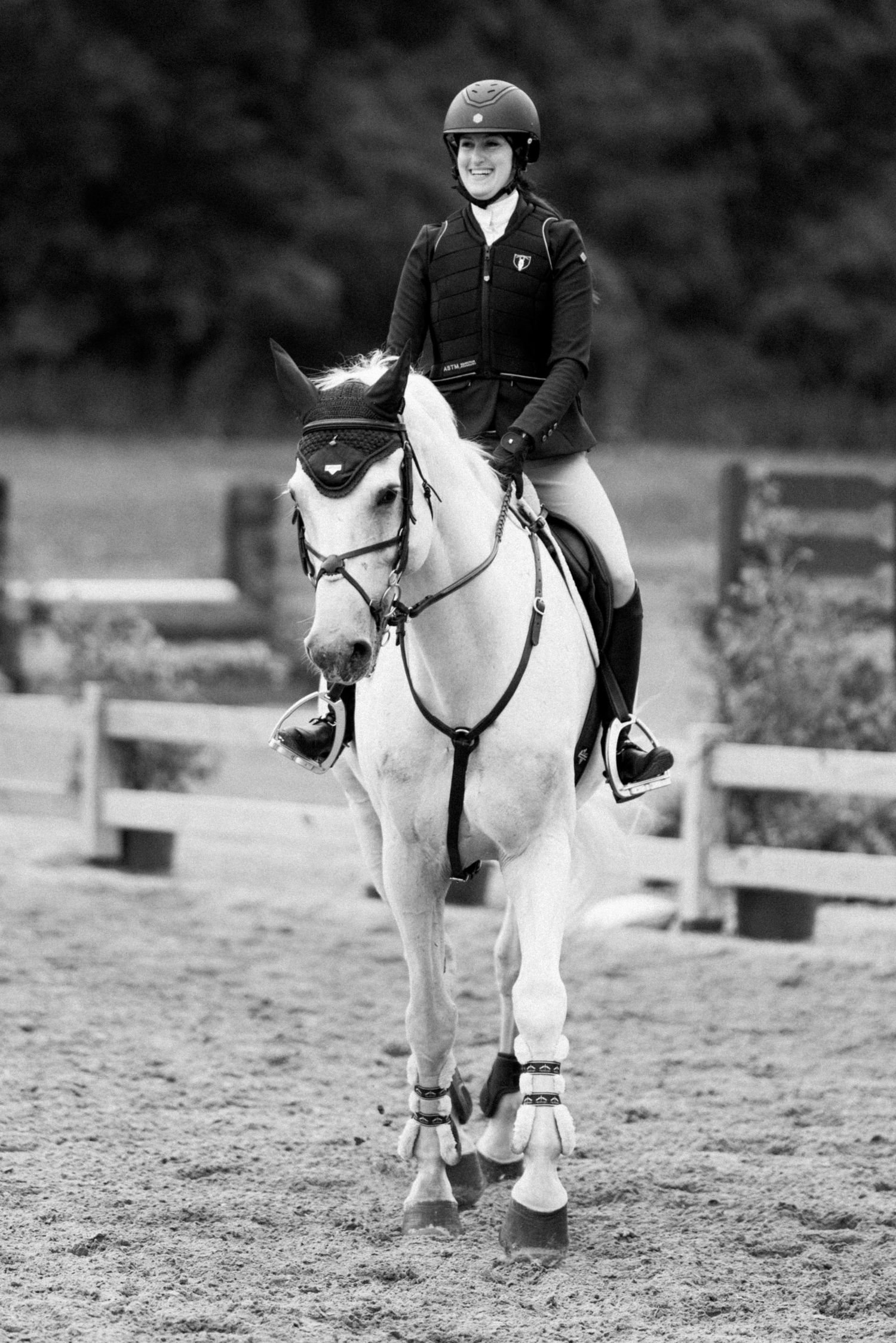 Black and white photo of a smiling equestrian rider in helmet atop a white horse in a sand arena.