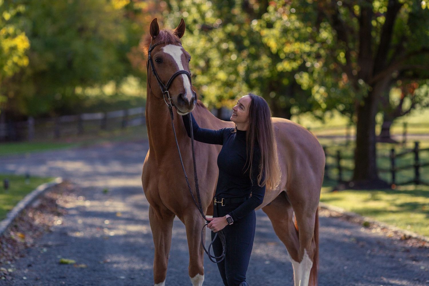 A woman in black attire stands beside a chestnut horse on a tree-lined path in a lush green setting.
