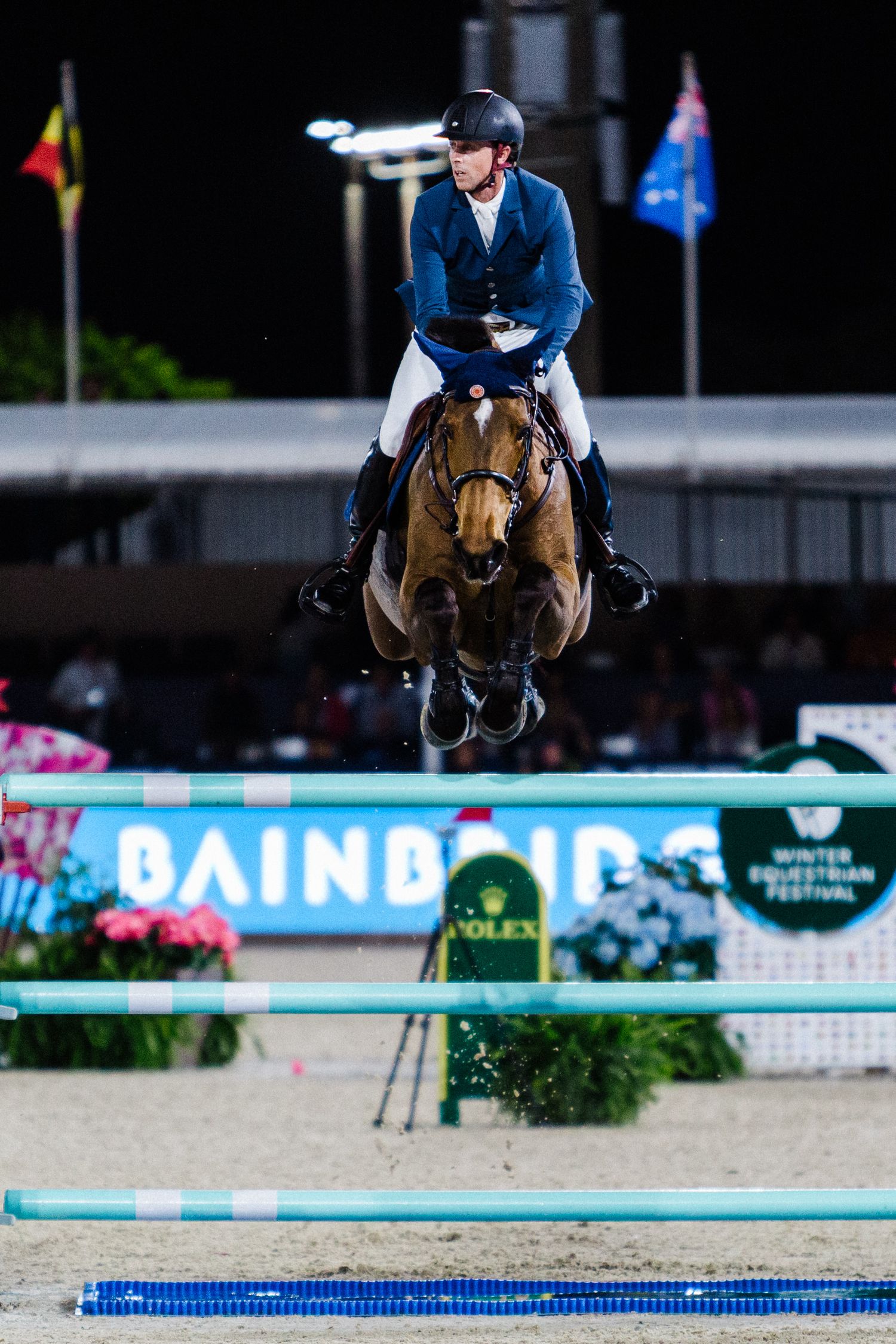 Equestrian rider on brown horse clearing a blue and white jump rail during competition at outdoor arena.