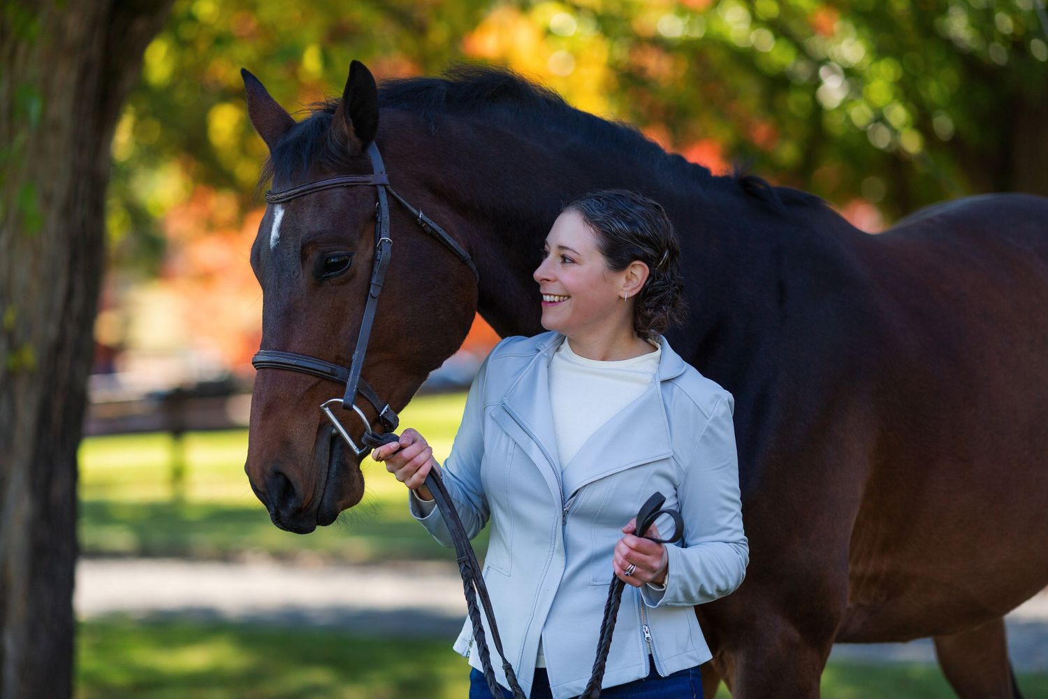 A person in a light blue jacket shares a tender moment with a dark brown horse in an autumn setting.