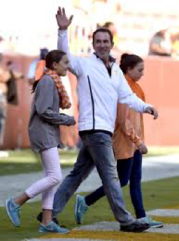 A man in a white jacket waves while walking on a football field with two women in orange and gray.
