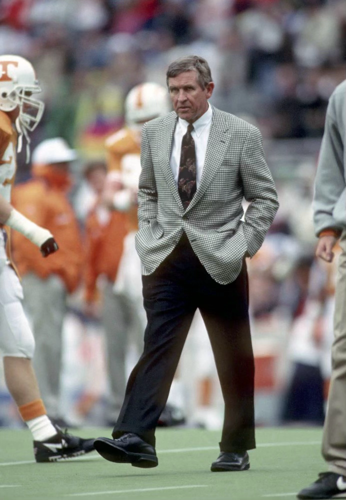 Football coach in gray sport coat and dark pants kneels on sideline during Tennessee Volunteers game.