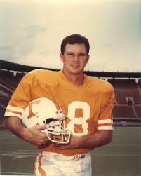 College football player in yellow jersey number 8 holds helmet on stadium field in vintage team photo.