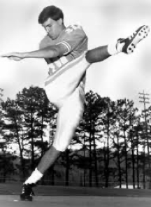 Black and white photo of a male athlete in uniform performing a high kick on an outdoor field with trees in background.