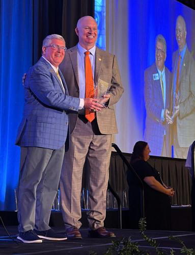 Two men share an award on stage at a formal event, one holding a glass trophy, with a large screen behind them.