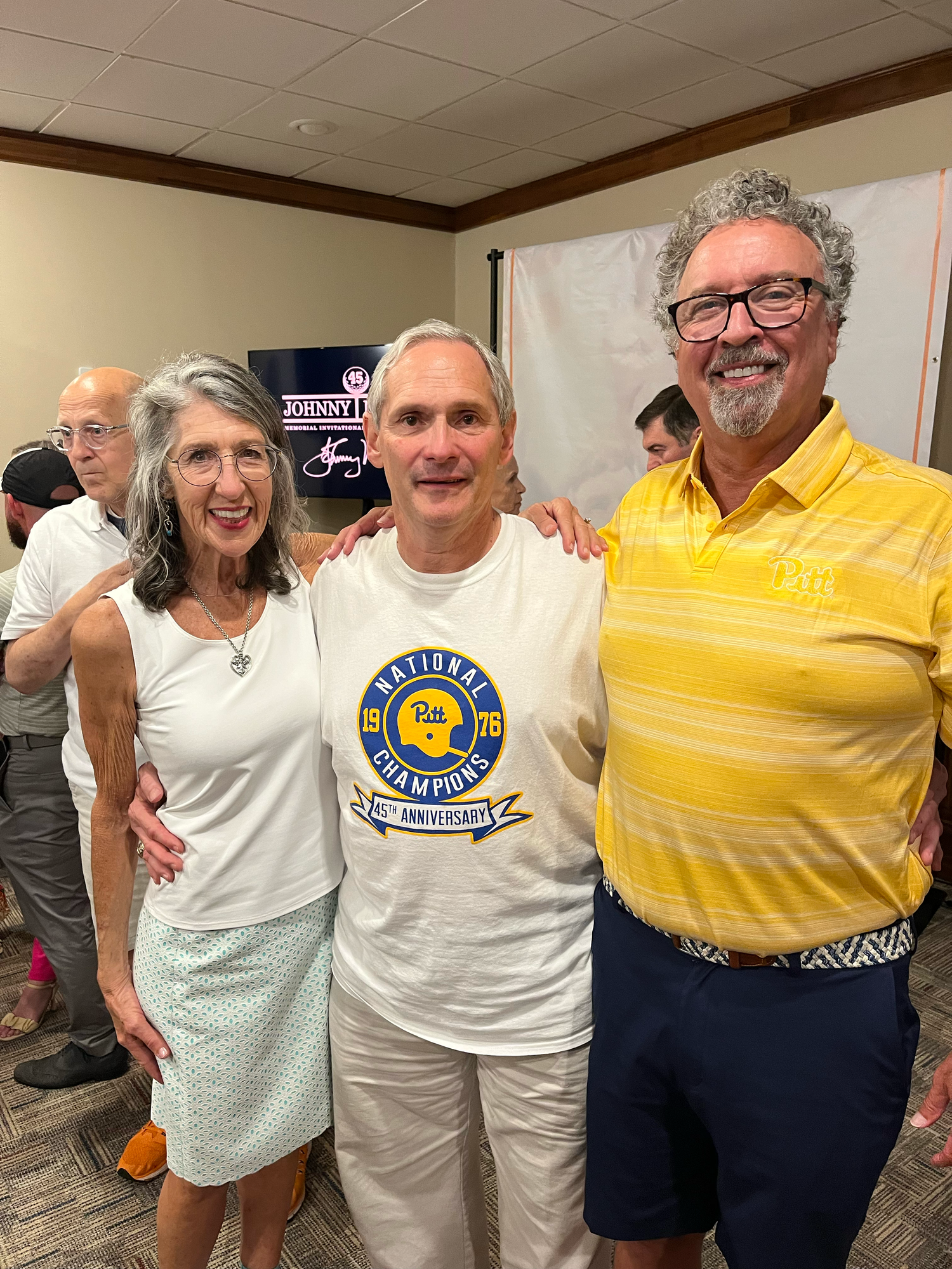 Three smiling adults pose together at a casual indoor event, center person wearing a National Champions anniversary t-shirt.