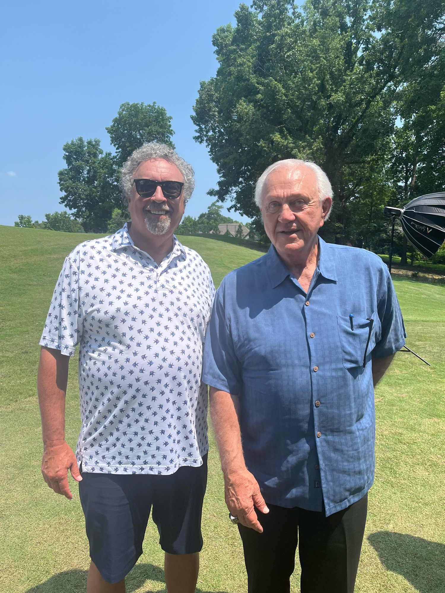 Two older men smiling together outdoors on a sunny golf course with lush green trees in the background.