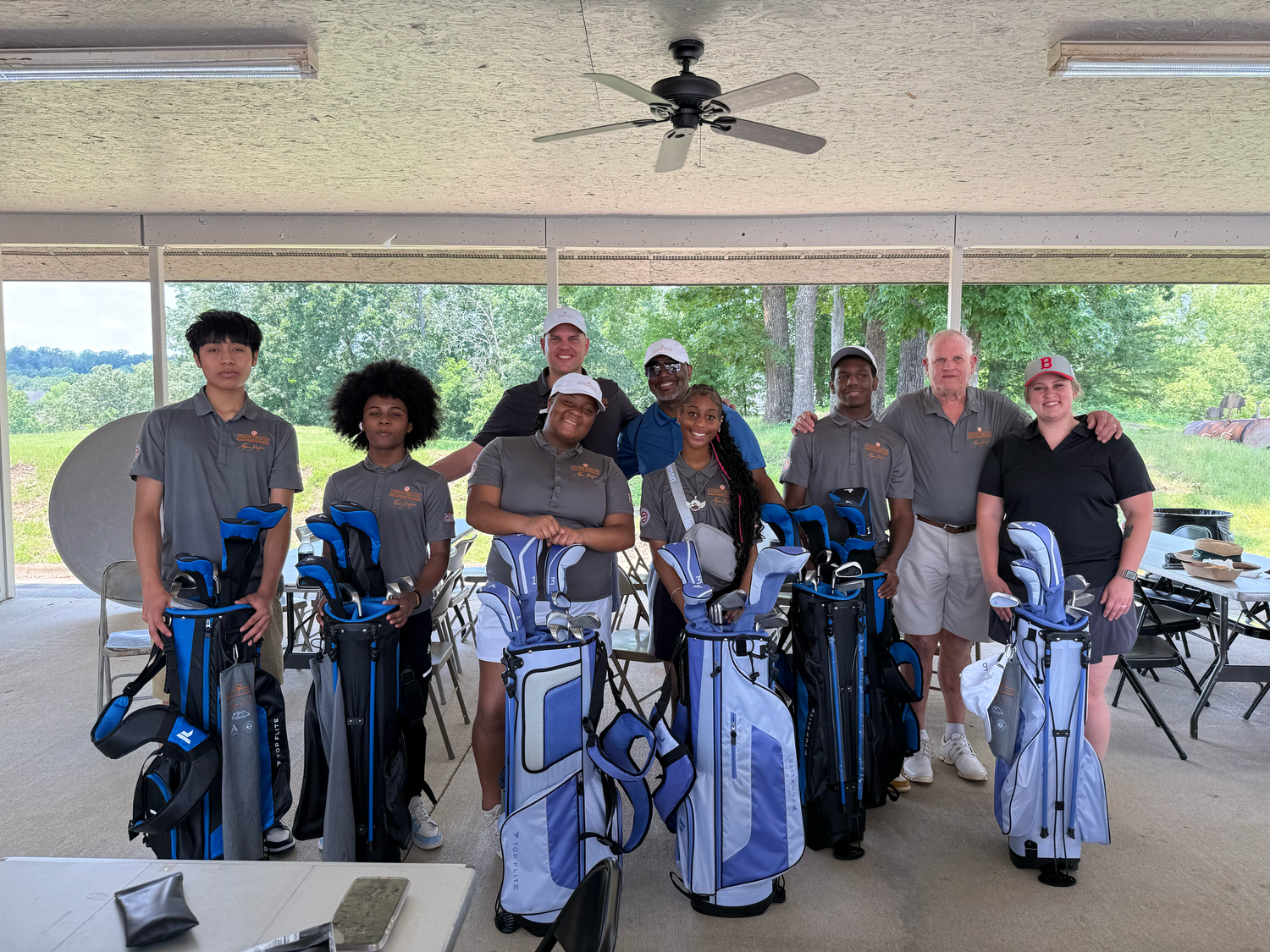 A group of young golfers and coaches pose with blue and gray golf bags under a covered pavilion outdoors.