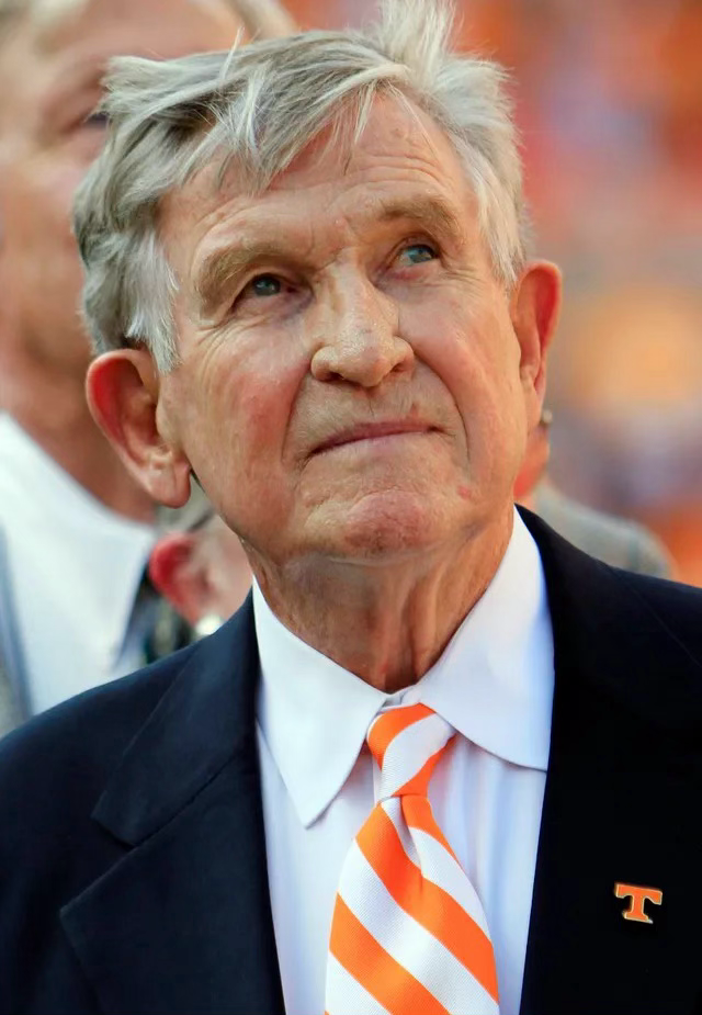 A person wearing a white dress shirt and orange striped tie with a University of Tennessee logo pin.