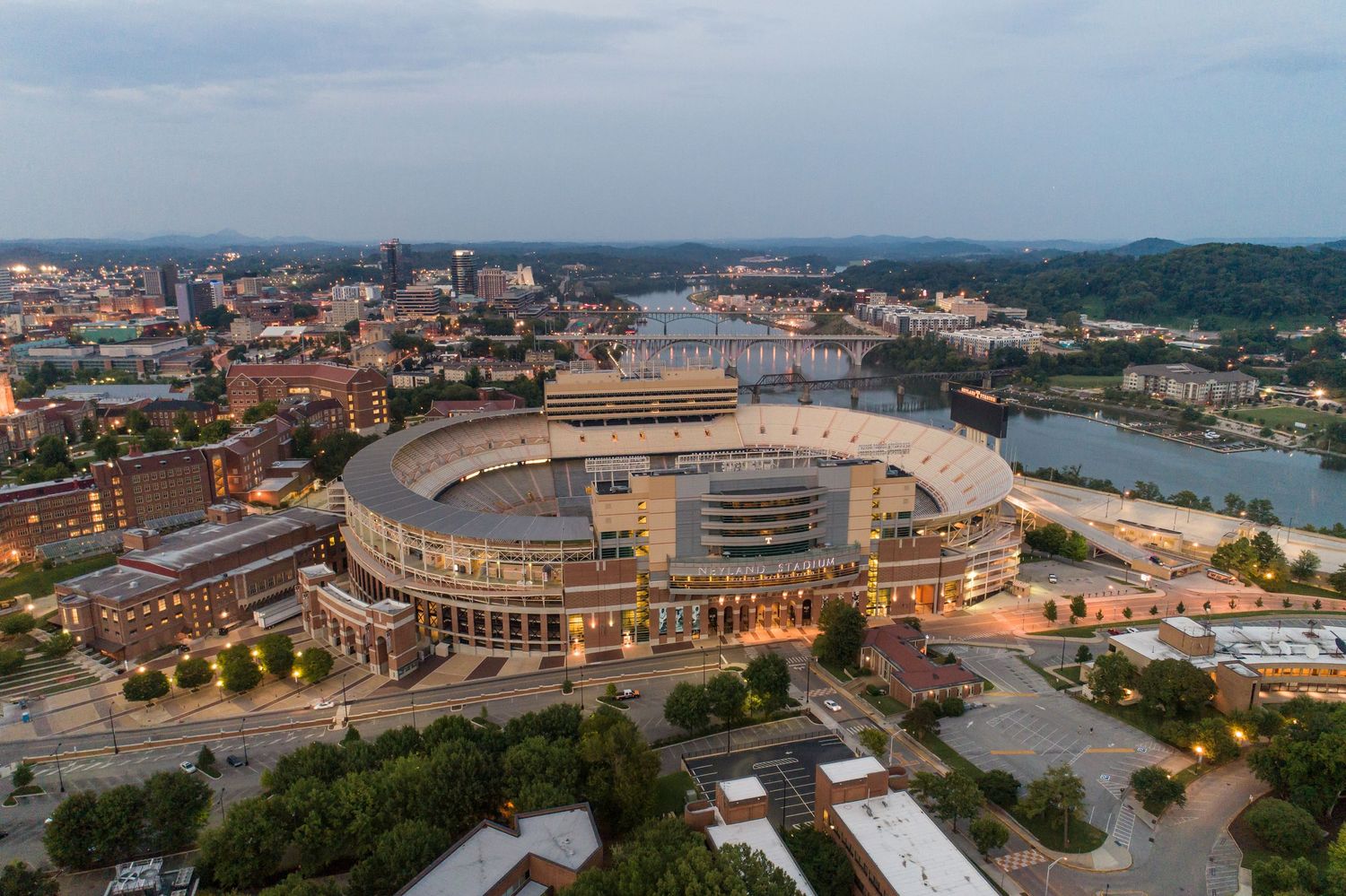 Aerial twilight view of Neyland Stadium in Knoxville, Tennessee, beside the Tennessee River with city skyline.