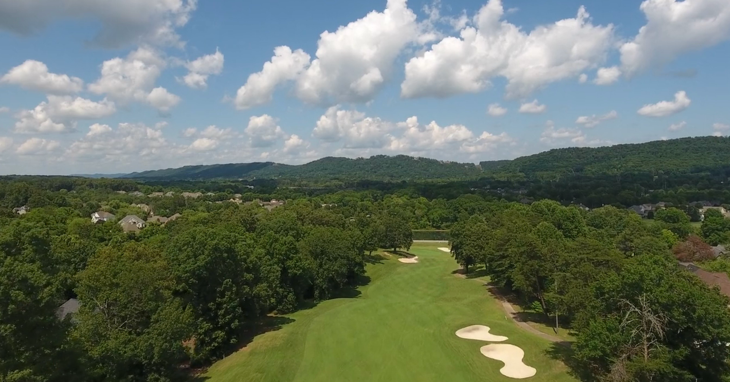 Aerial view of a lush green golf course with sand bunkers, surrounded by trees and rolling hills under a partly cloudy sky.