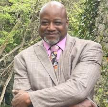 Man in gray suit and pink tie smiling with arms crossed outdoors among green trees.