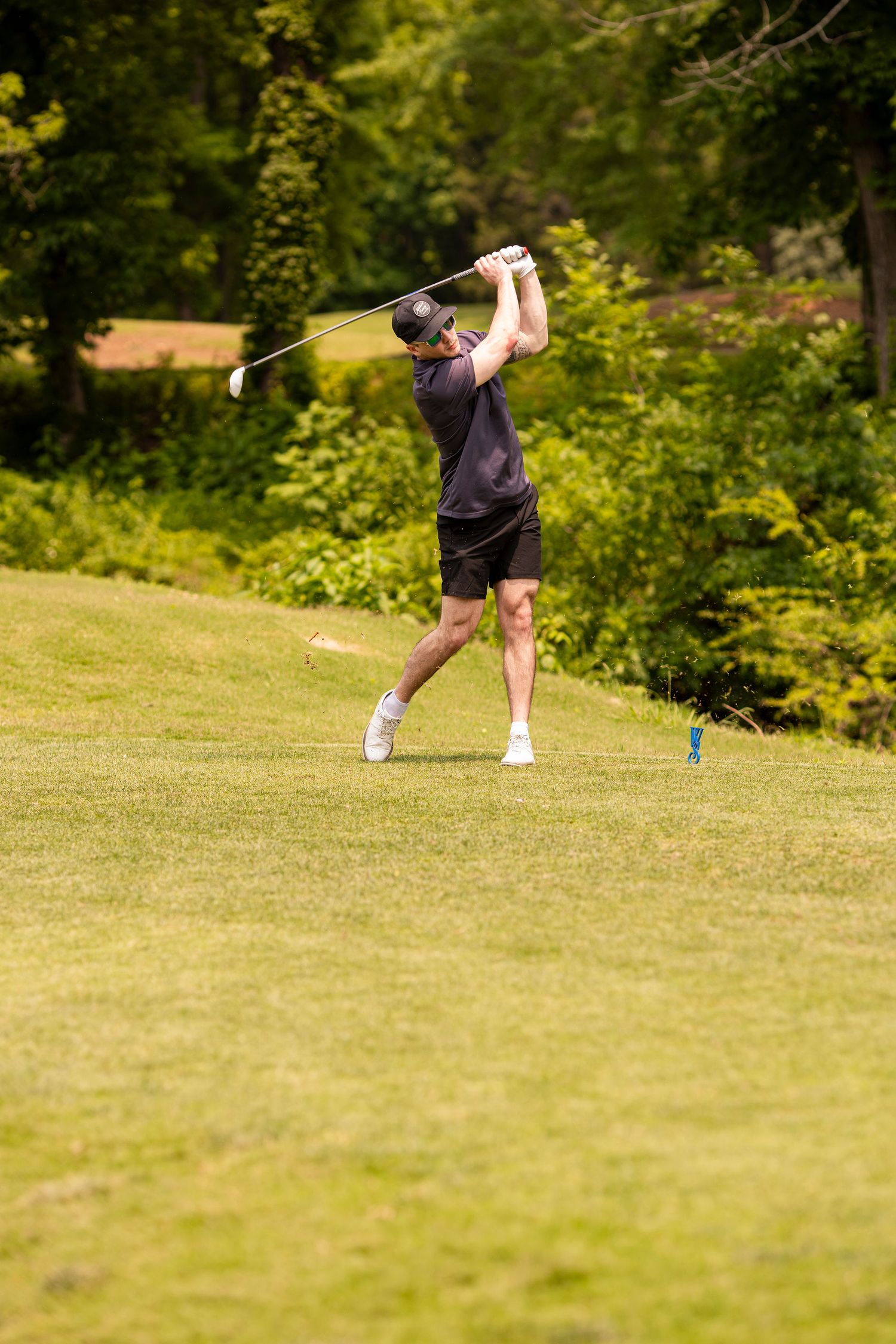 Multiple golfers taking shots on a lush green golf course with trees in the background.