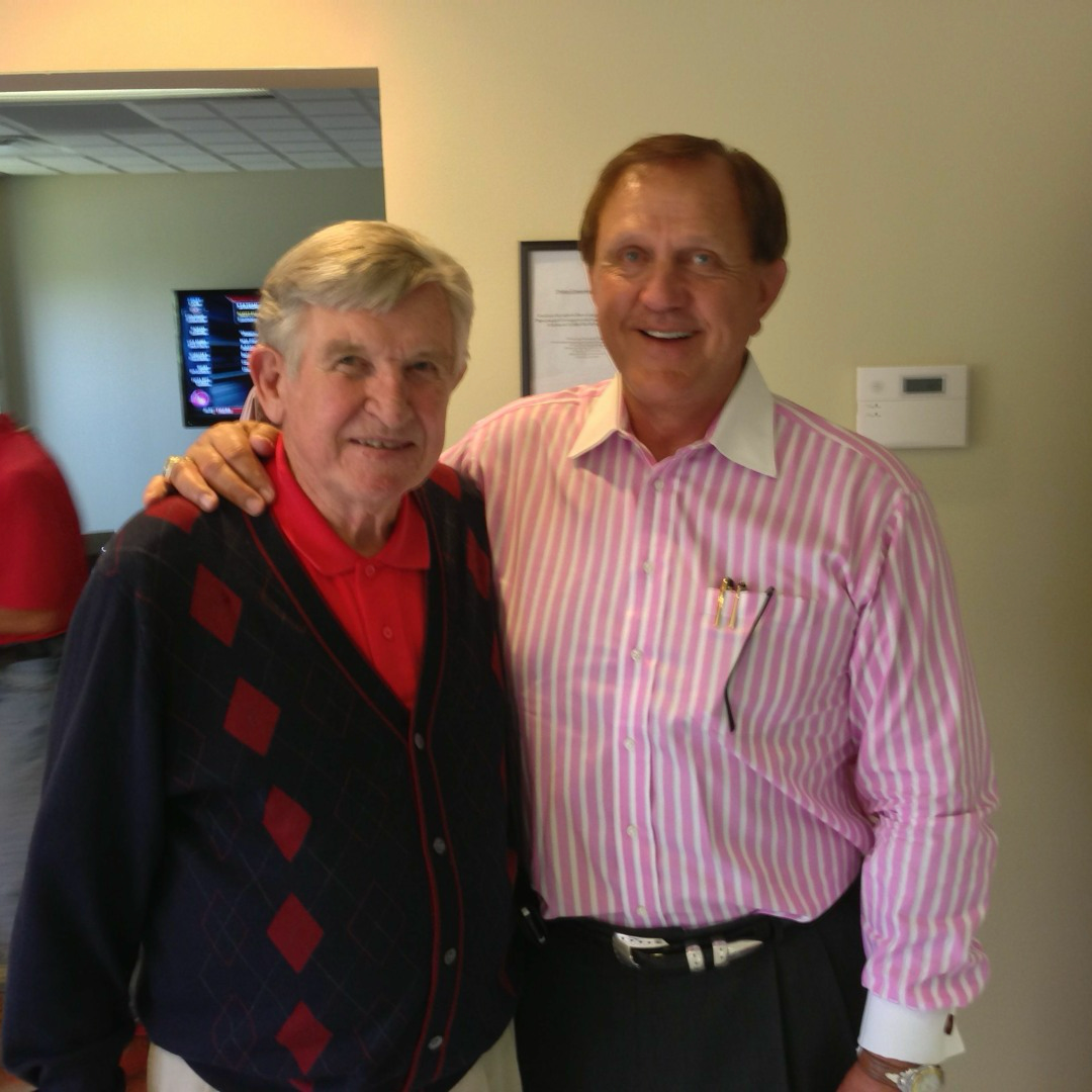 Two elderly men smiling and posing together, one in a red argyle cardigan and one in a pink striped shirt.
