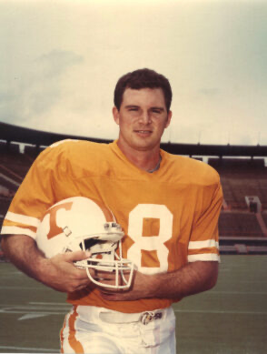 College football player in orange jersey number 8 holds helmet on stadium field.