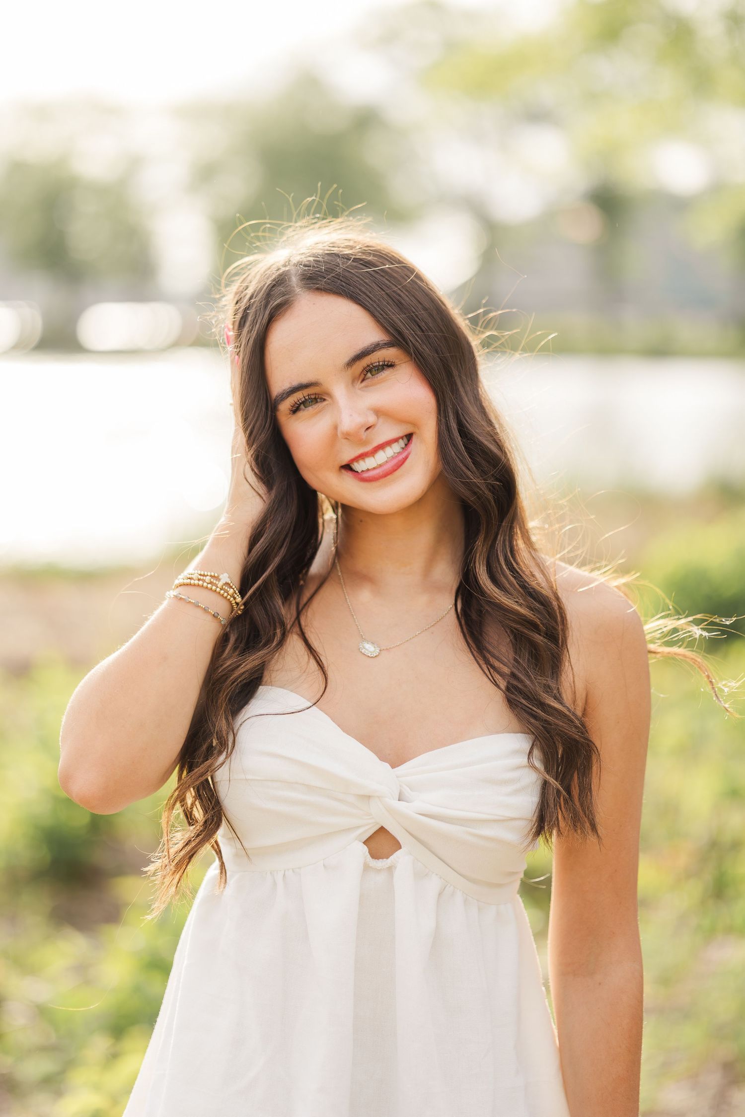 Young woman in white sundress smiling outdoors near water, wearing gold bracelets and delicate necklace.