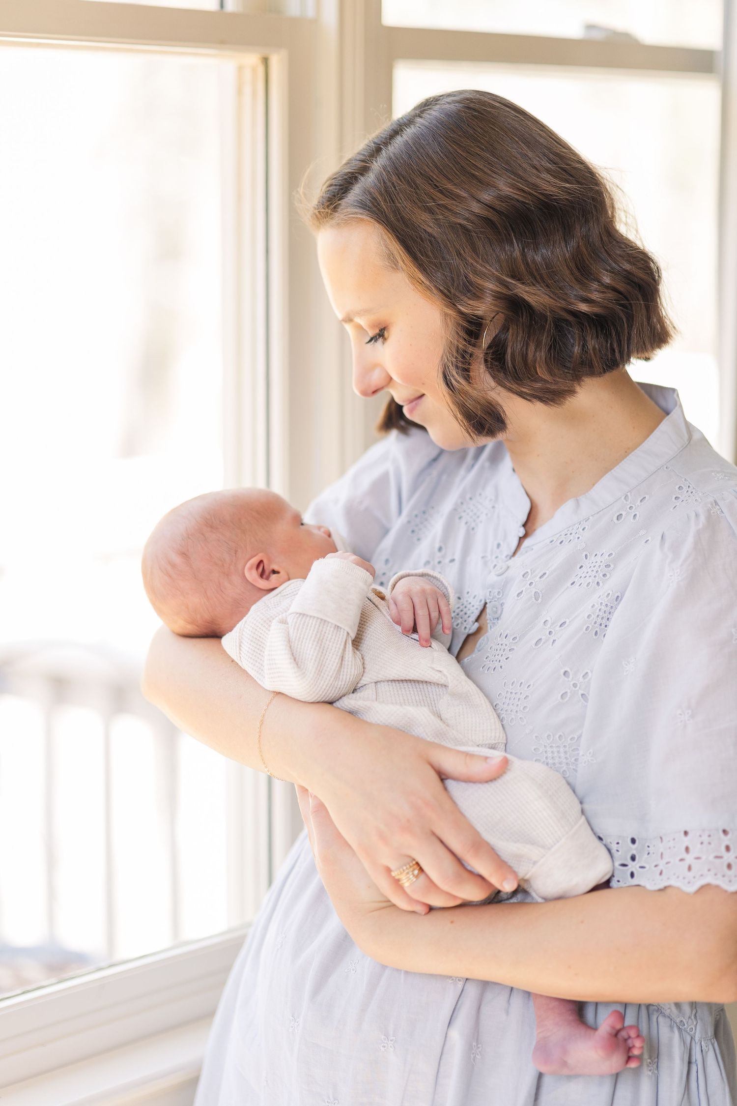 A mother in a light blue dress lovingly gazes at her newborn baby held gently in her arms by a bright window.