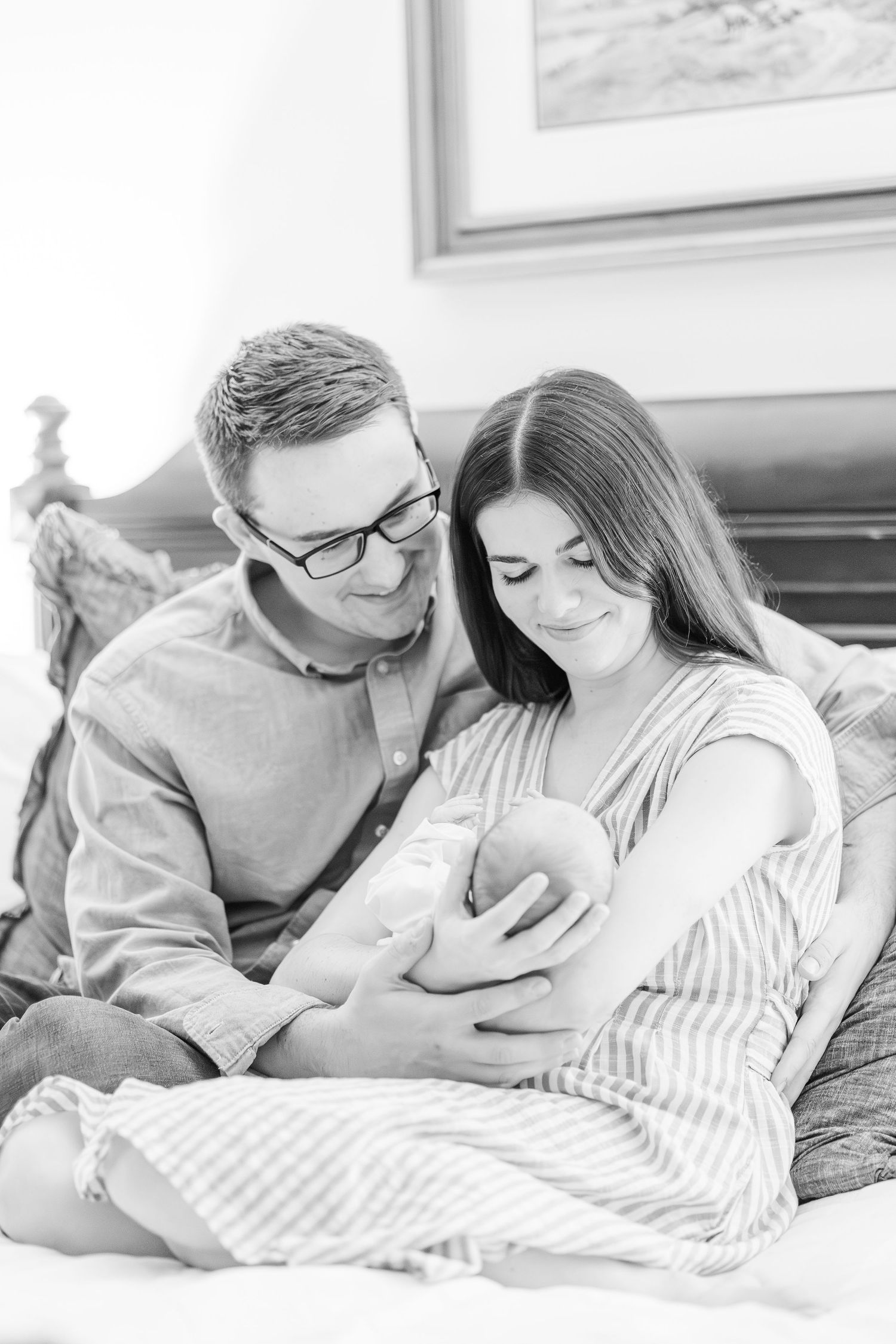 Black and white photo of smiling parents sitting on bed, lovingly cradling their newborn baby together.