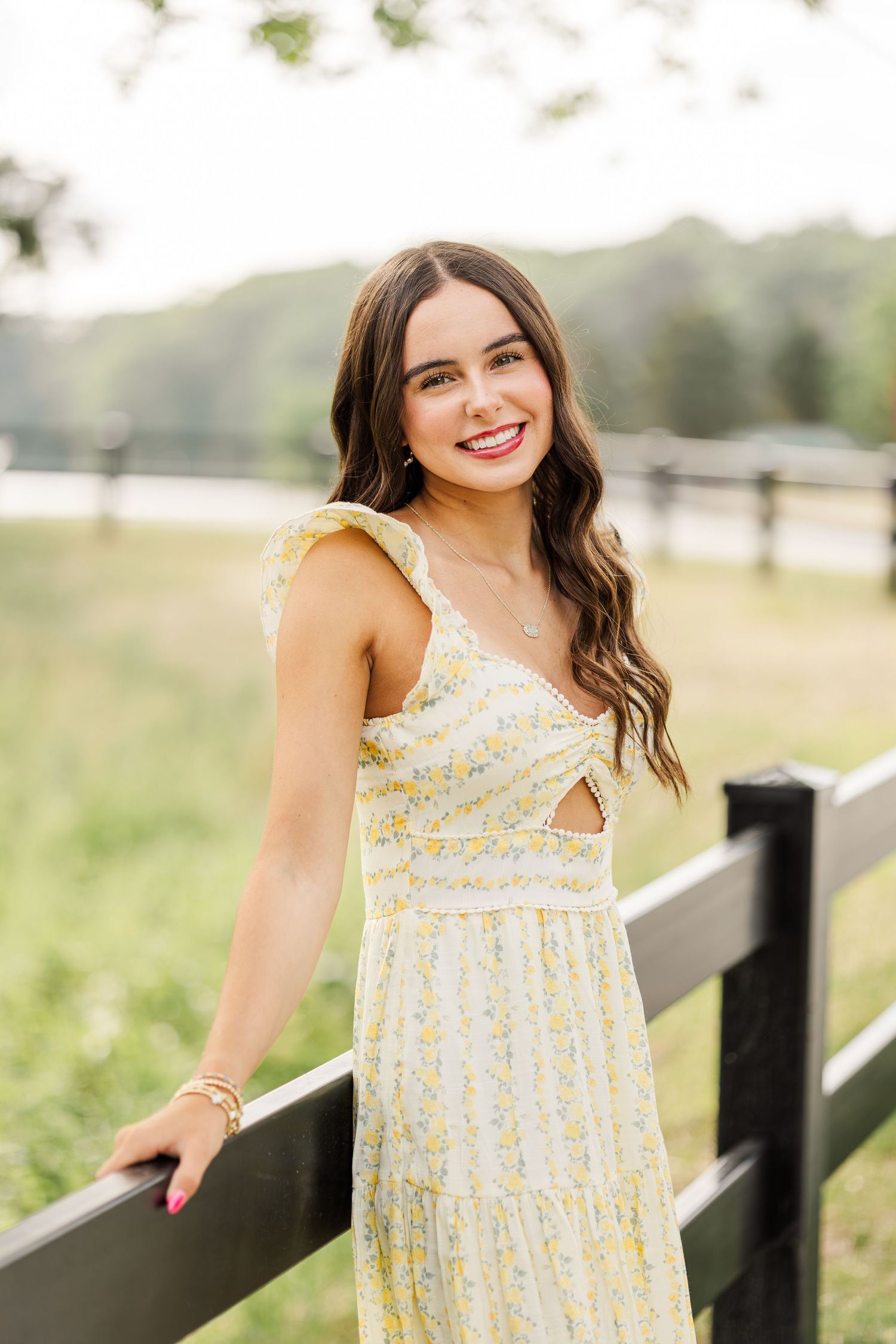 Young woman in yellow floral midi dress smiling by a black wooden fence in a lush green outdoor setting.