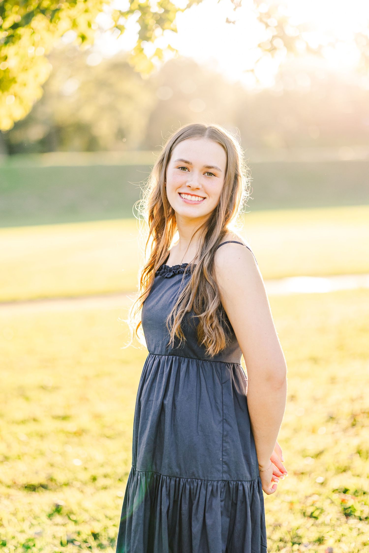 Smiling teenage girl in navy dress standing in sunlit park with golden hour light filtering through trees.