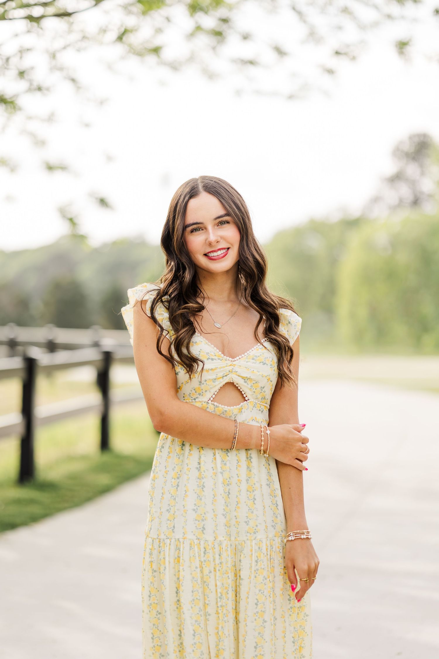Young woman in yellow floral midi dress smiling outdoors on a scenic path with lush green trees.