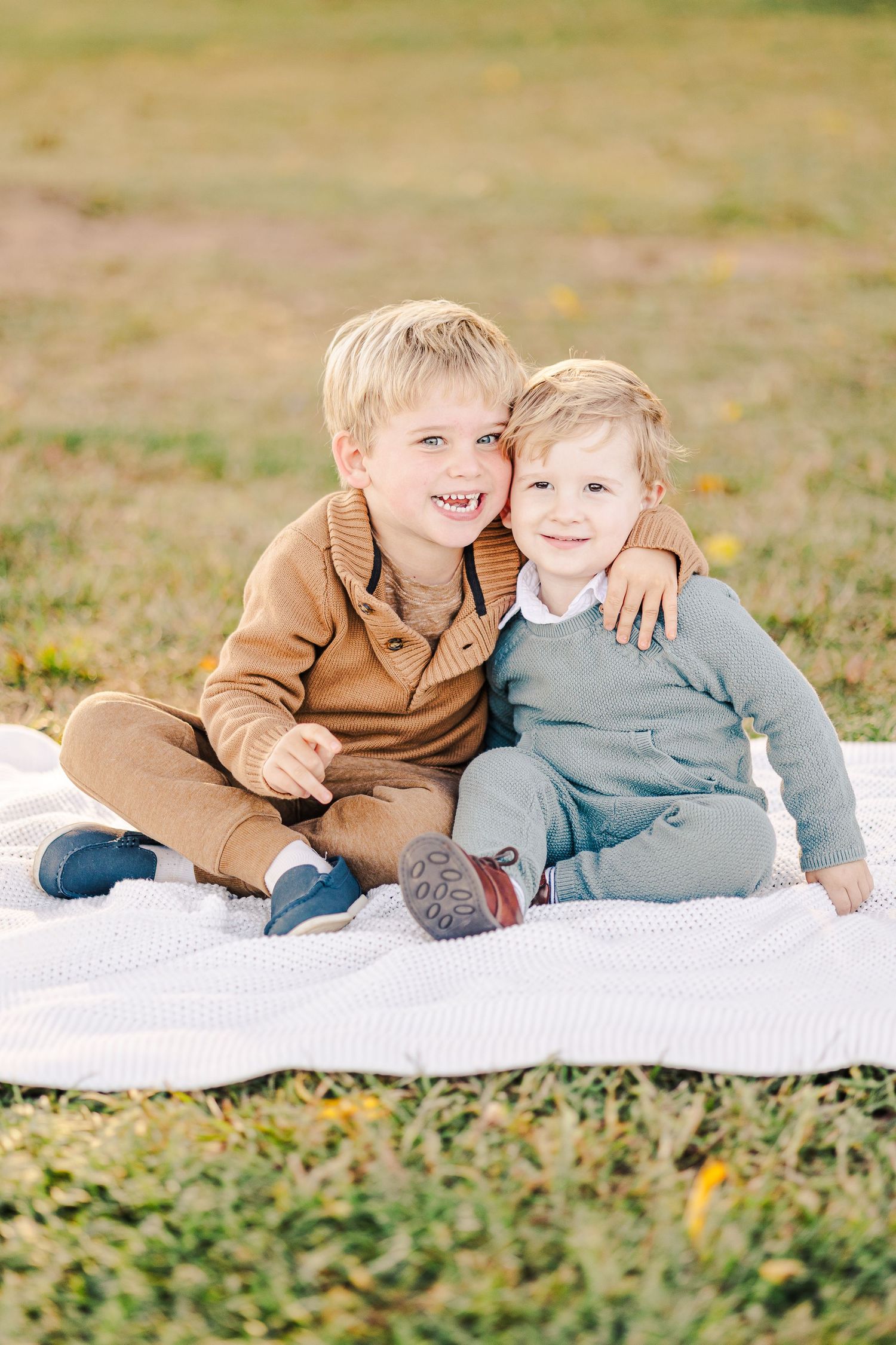 Two young blonde brothers sitting together on a white blanket outdoors, smiling and hugging in autumn.