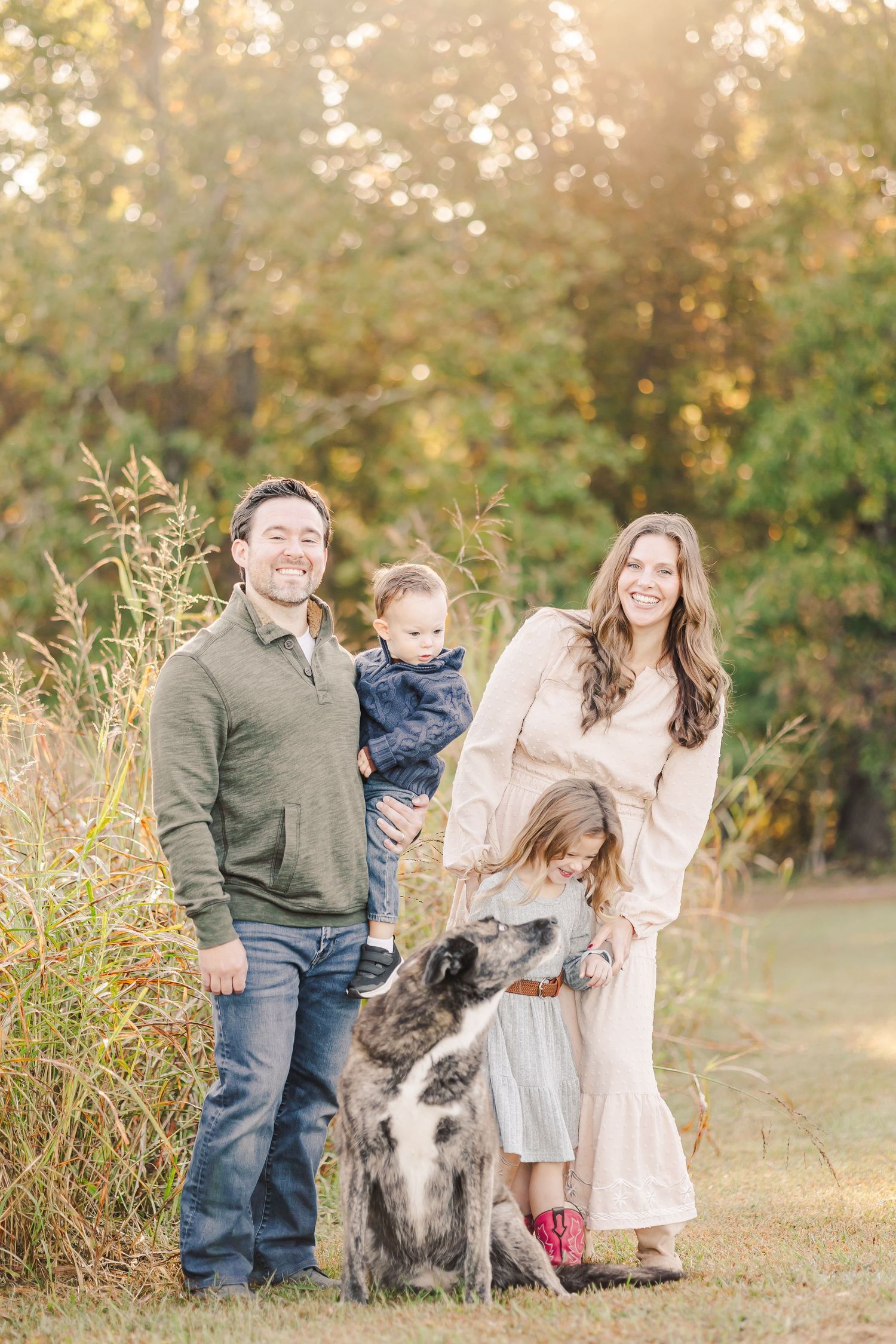 Family of four with dog posing in autumn field, parents holding children among golden fall foliage.