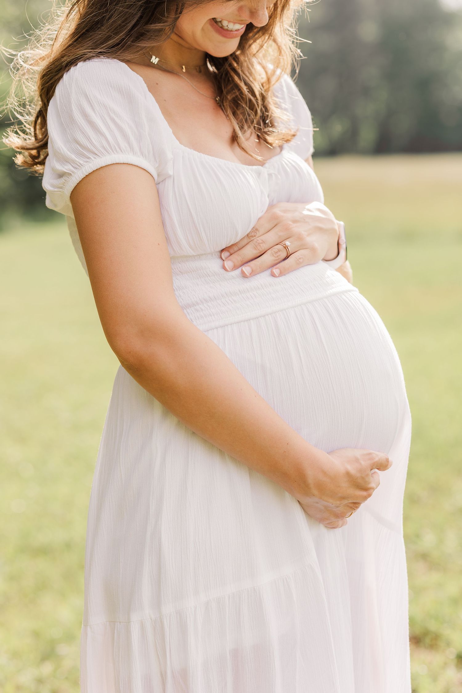 Pregnant woman in white dress cradling baby bump outdoors in a sunlit field during maternity photo session.