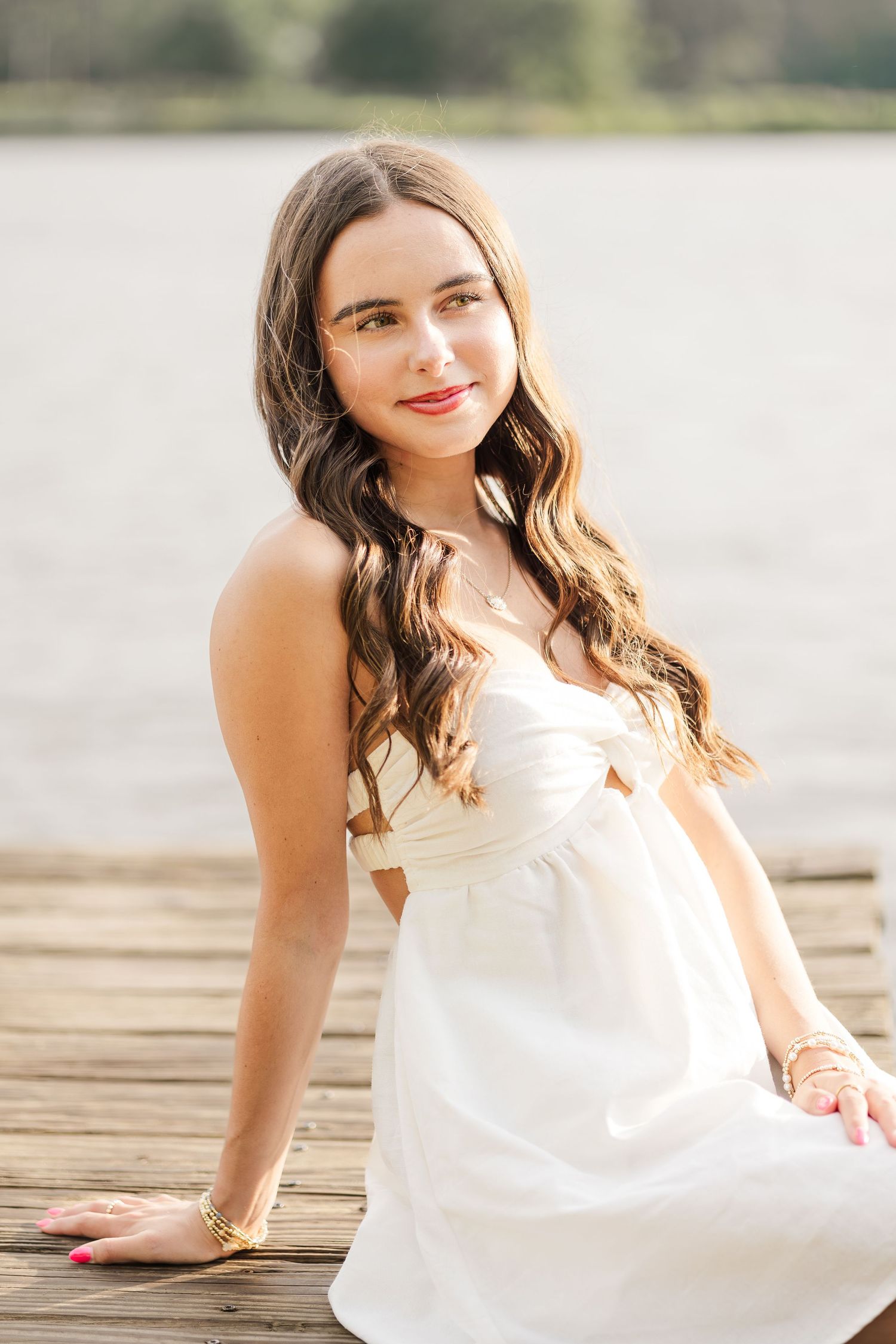 Young woman in white dress sitting on wooden dock by lake, smiling with long wavy brown hair.