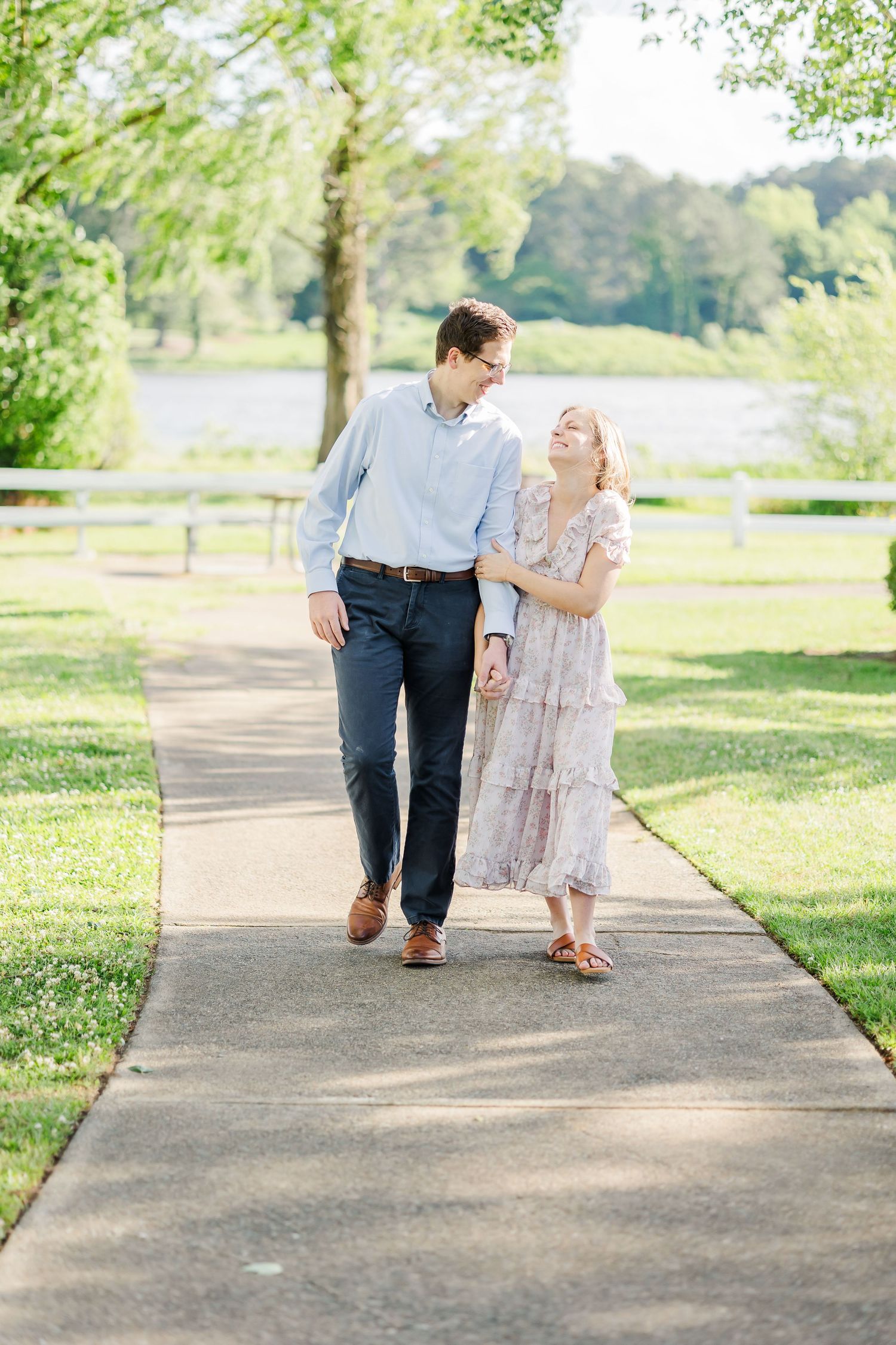 Engaged couple walks hand in hand along a sunny tree-lined path near a lake.