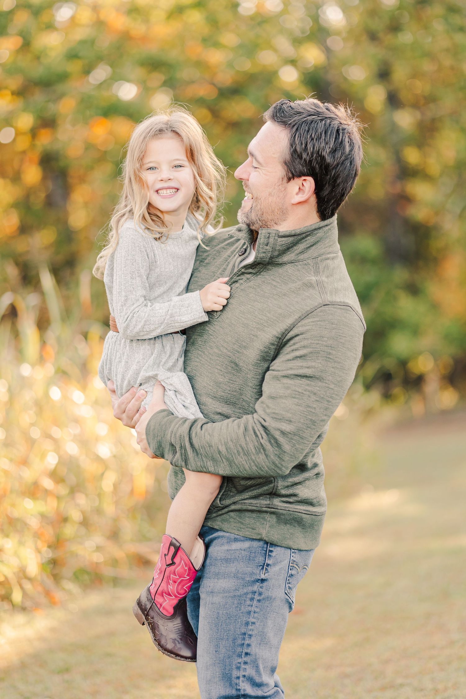 Father holds his laughing young daughter wearing cowboy boots during an autumn outdoor photo session.