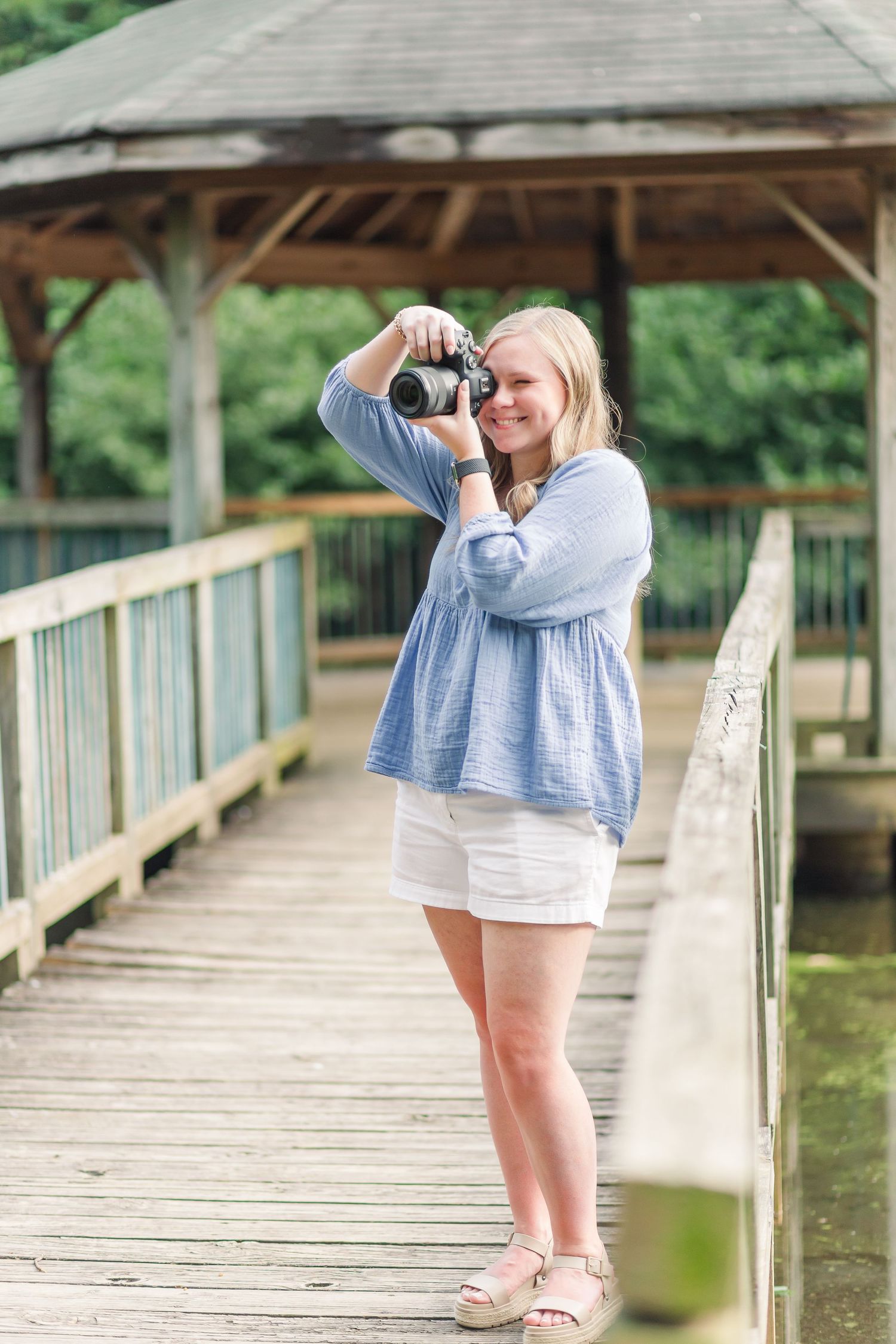 Smiling blonde woman photographing with mirrorless camera on wooden bridge dock in casual summer outfit.