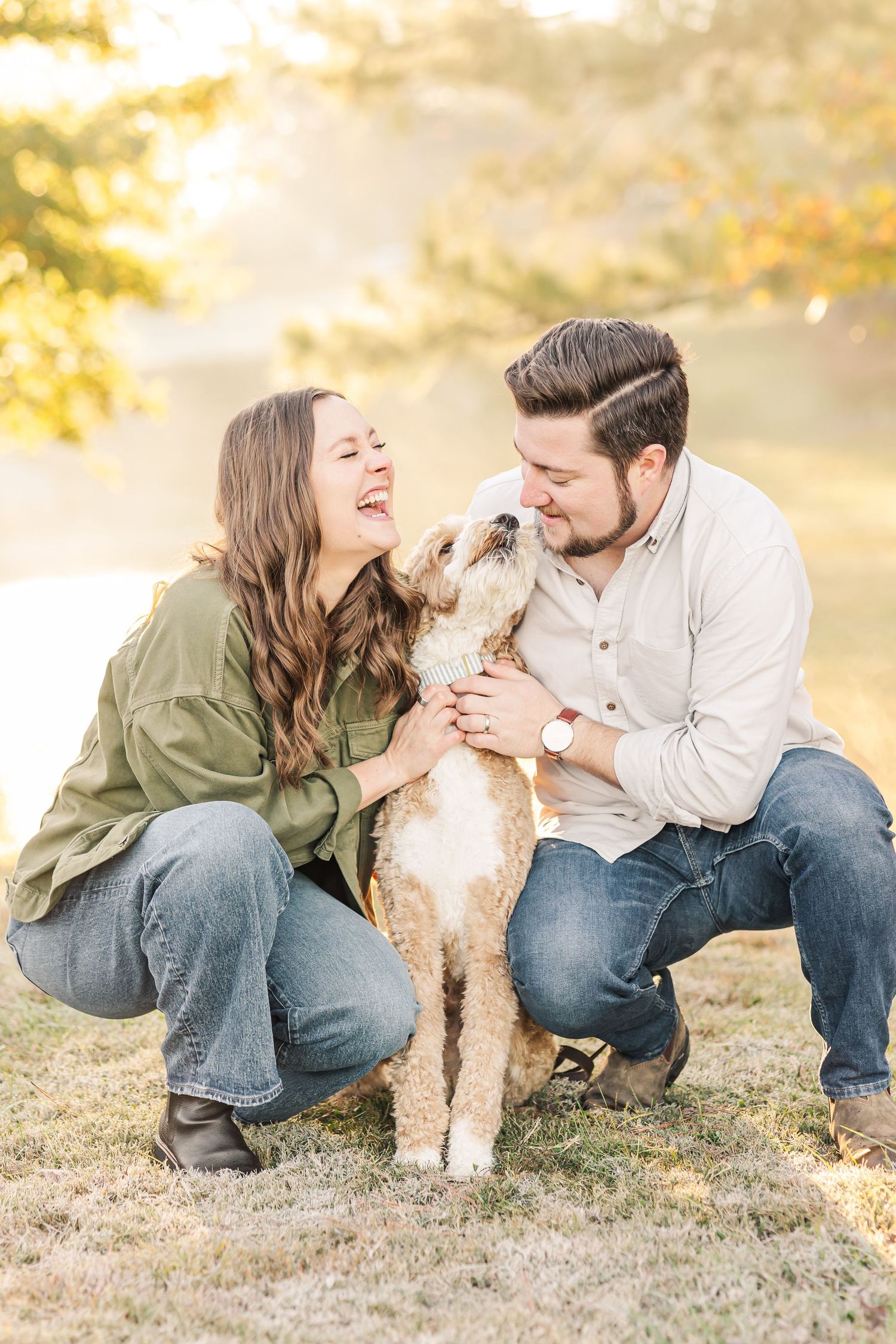 Couple crouching outdoors with their fluffy golden doodle during a golden hour engagement photo session.