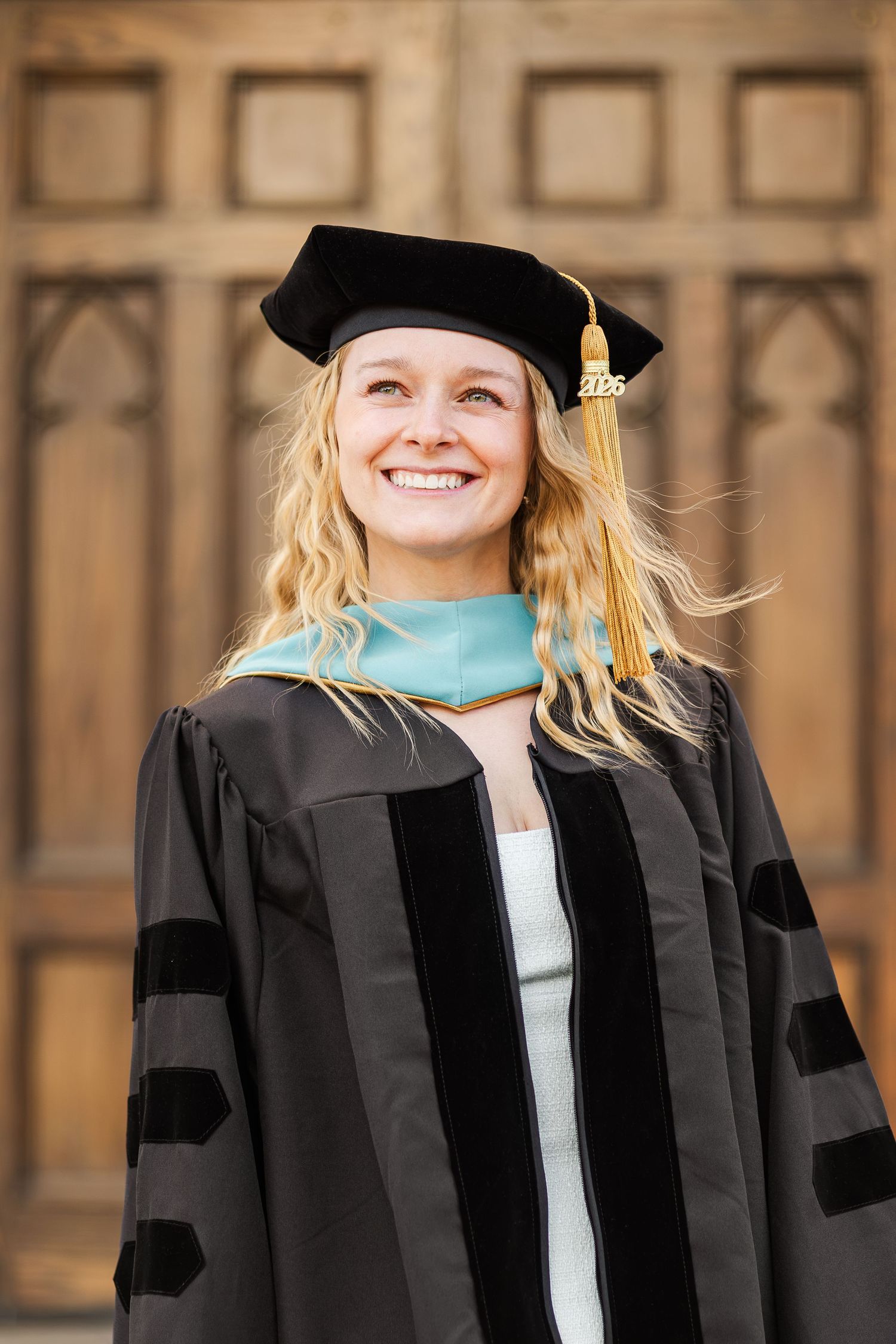 Smiling woman in black doctoral graduation gown and cap with gold tassel and teal hood, posing before ornate wooden doors.