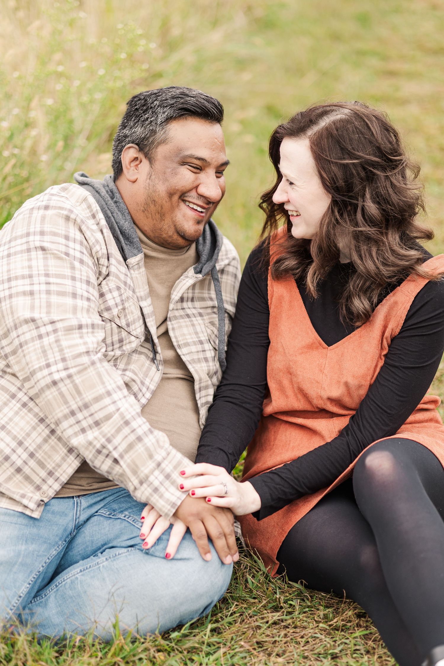 Happy couple sitting in grass holding hands, laughing together during outdoor photo session.