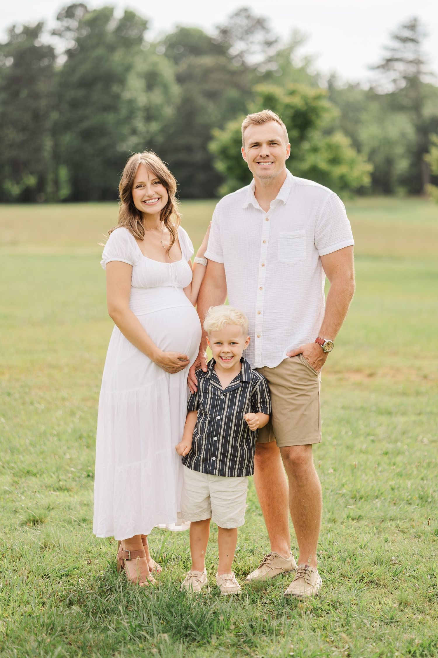 Pregnant woman in white dress poses with husband and young son in a sunny green field for a maternity photo.
