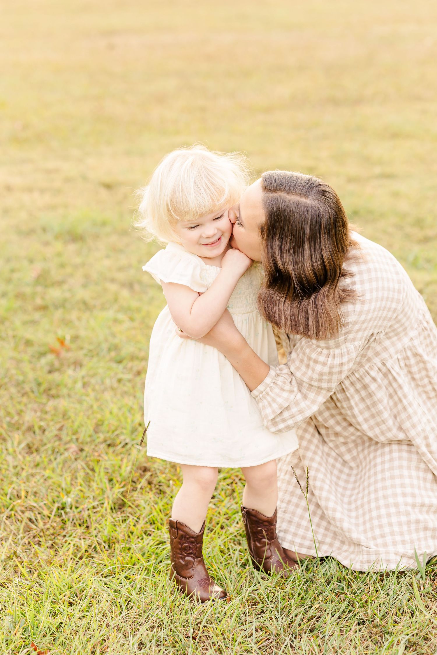 Mother kneeling in grass kisses her toddler daughter wearing brown cowboy boots outdoors.