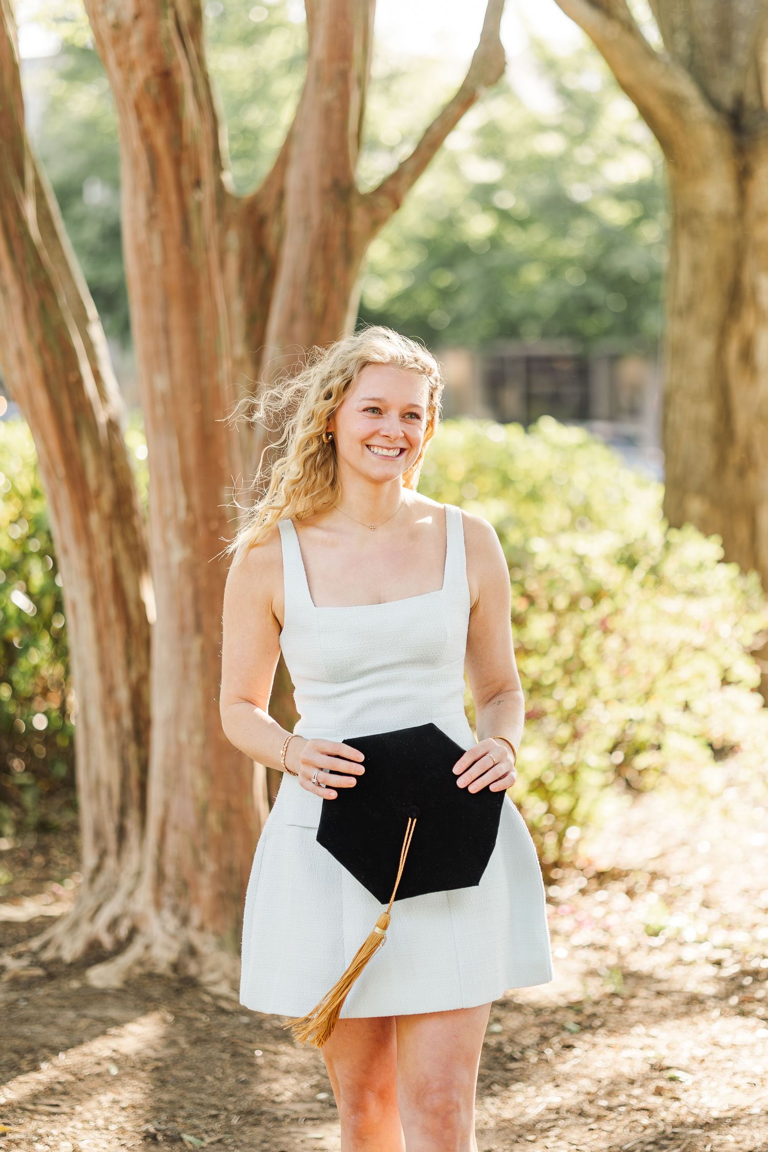 Smiling blonde woman in white dress holding graduation cap outdoors among trees on a sunny day.