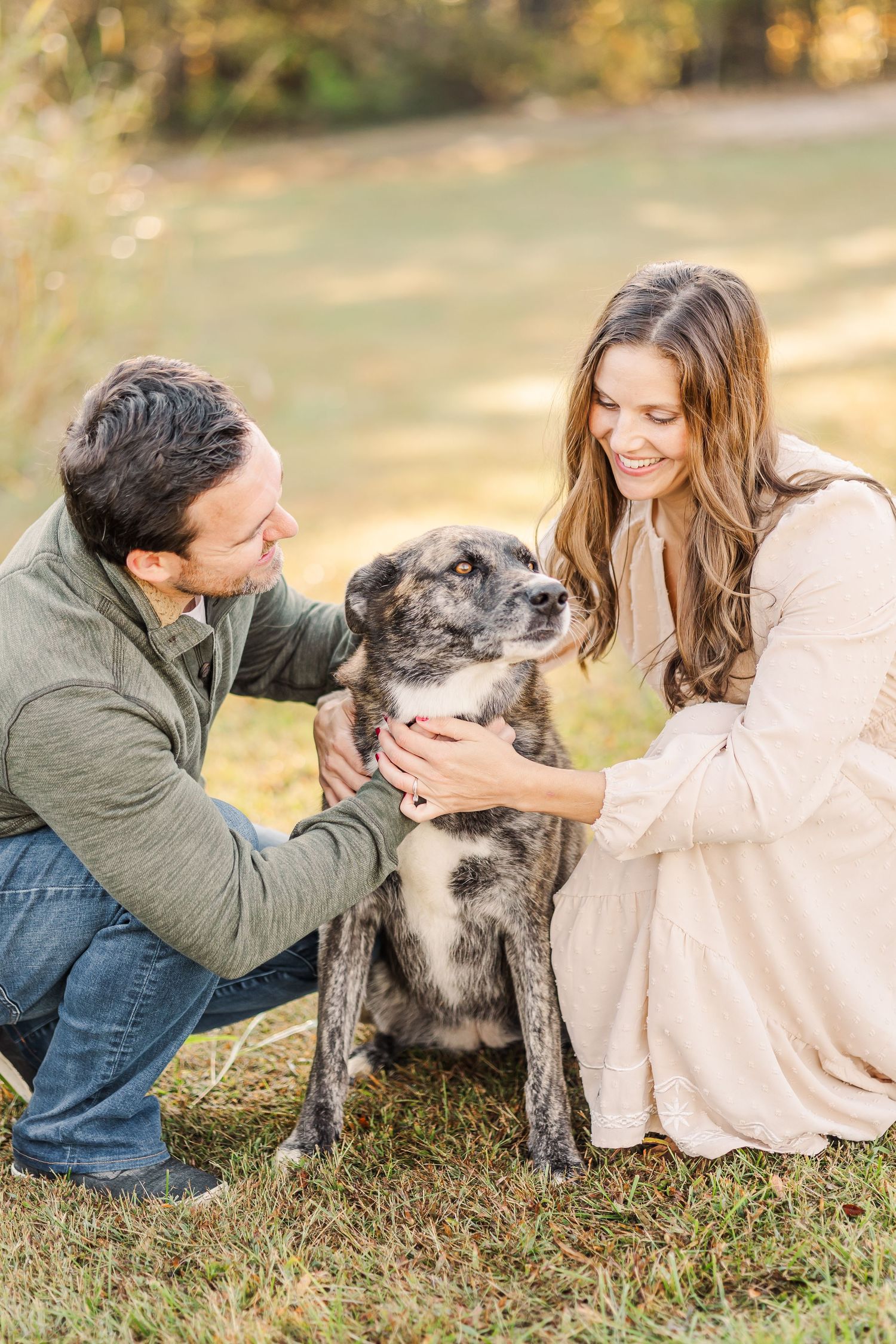 Couple crouching together petting their brindle dog in sunlit autumn park setting.